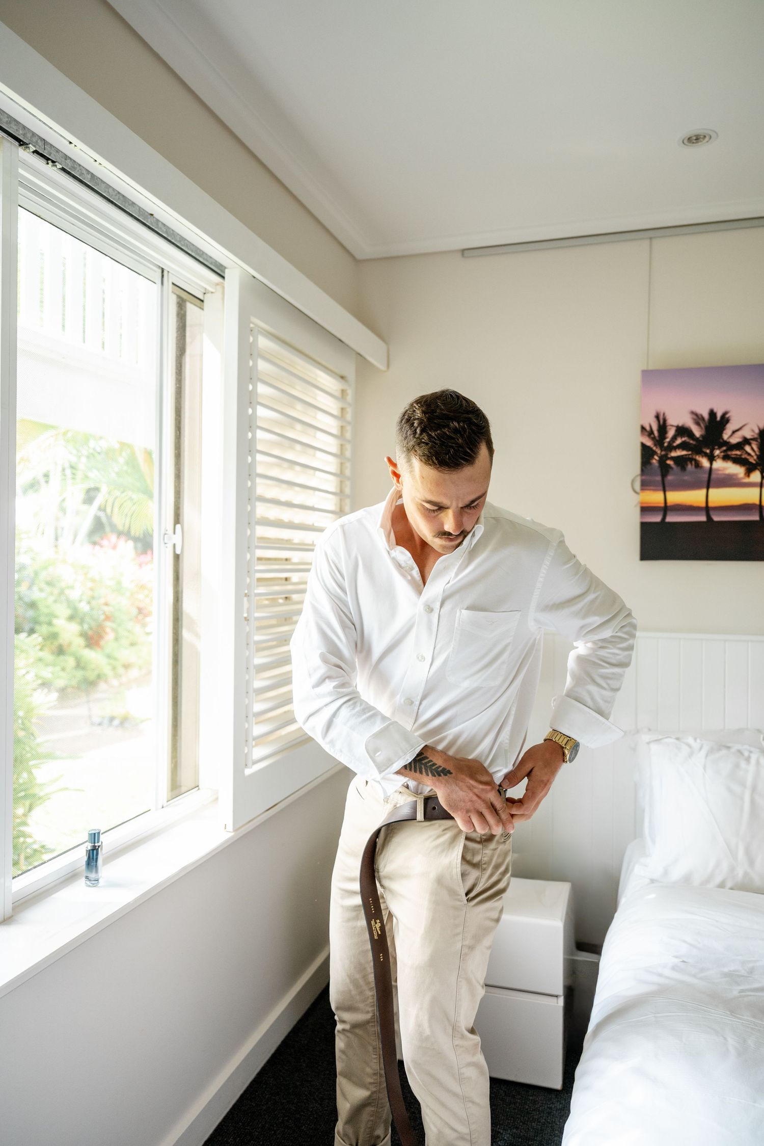 Groom in white shirt and khaki pants fastening his brown leather belt in a bright bedroom.