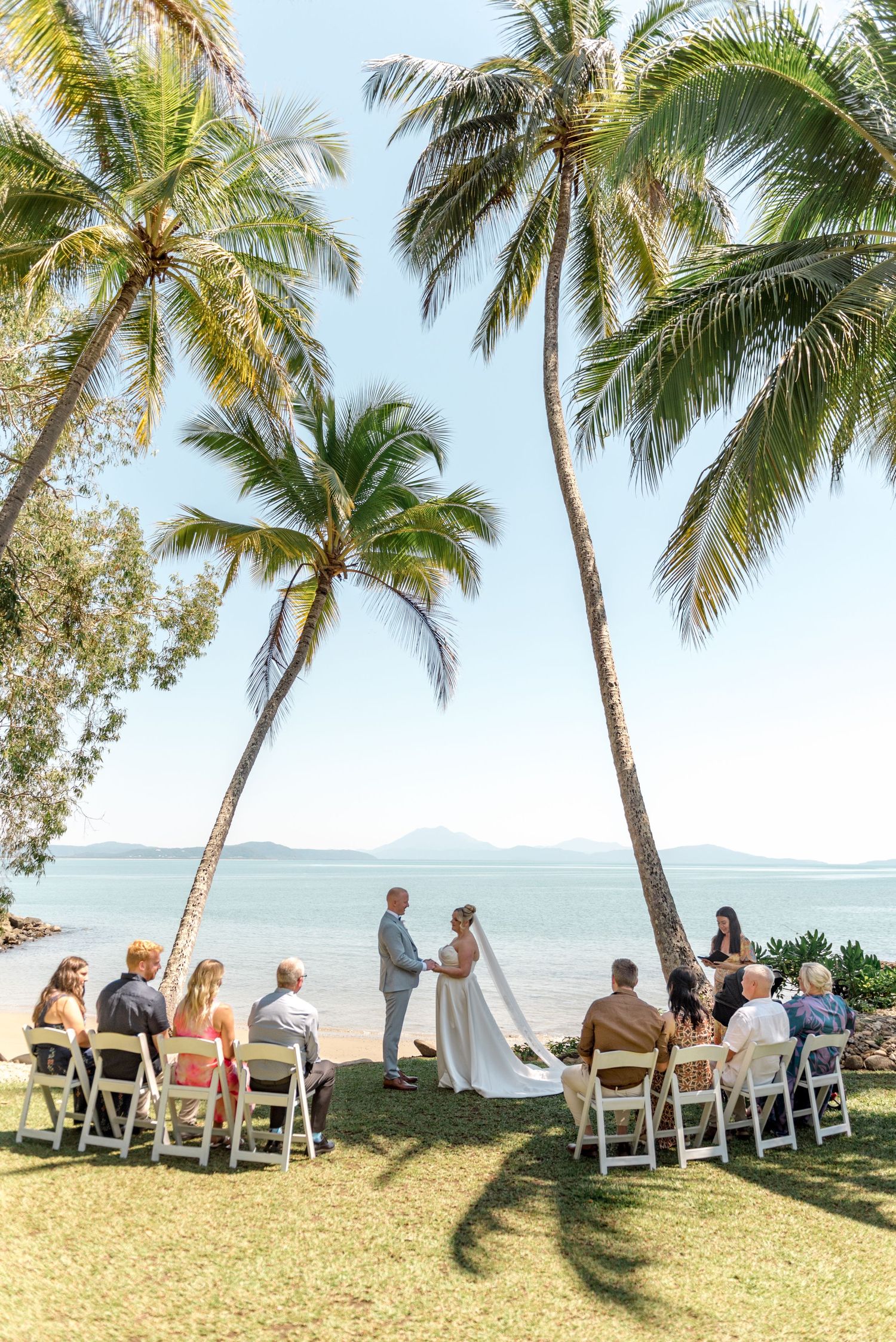 Tropical beach wedding ceremony under tall palm trees with ocean and mountain views in background.