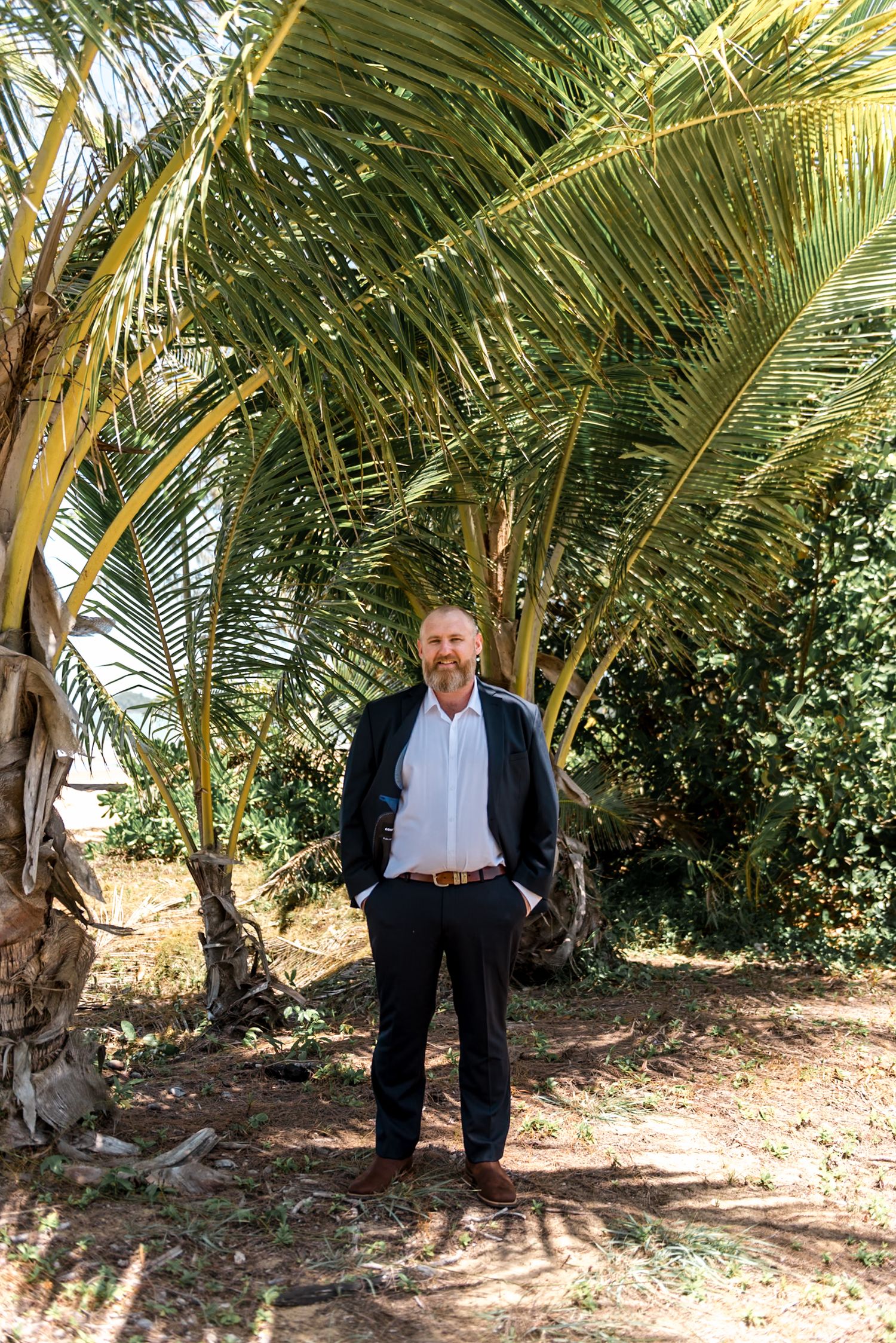 Smiling bearded man in a dark suit standing under large tropical palm trees outdoors.