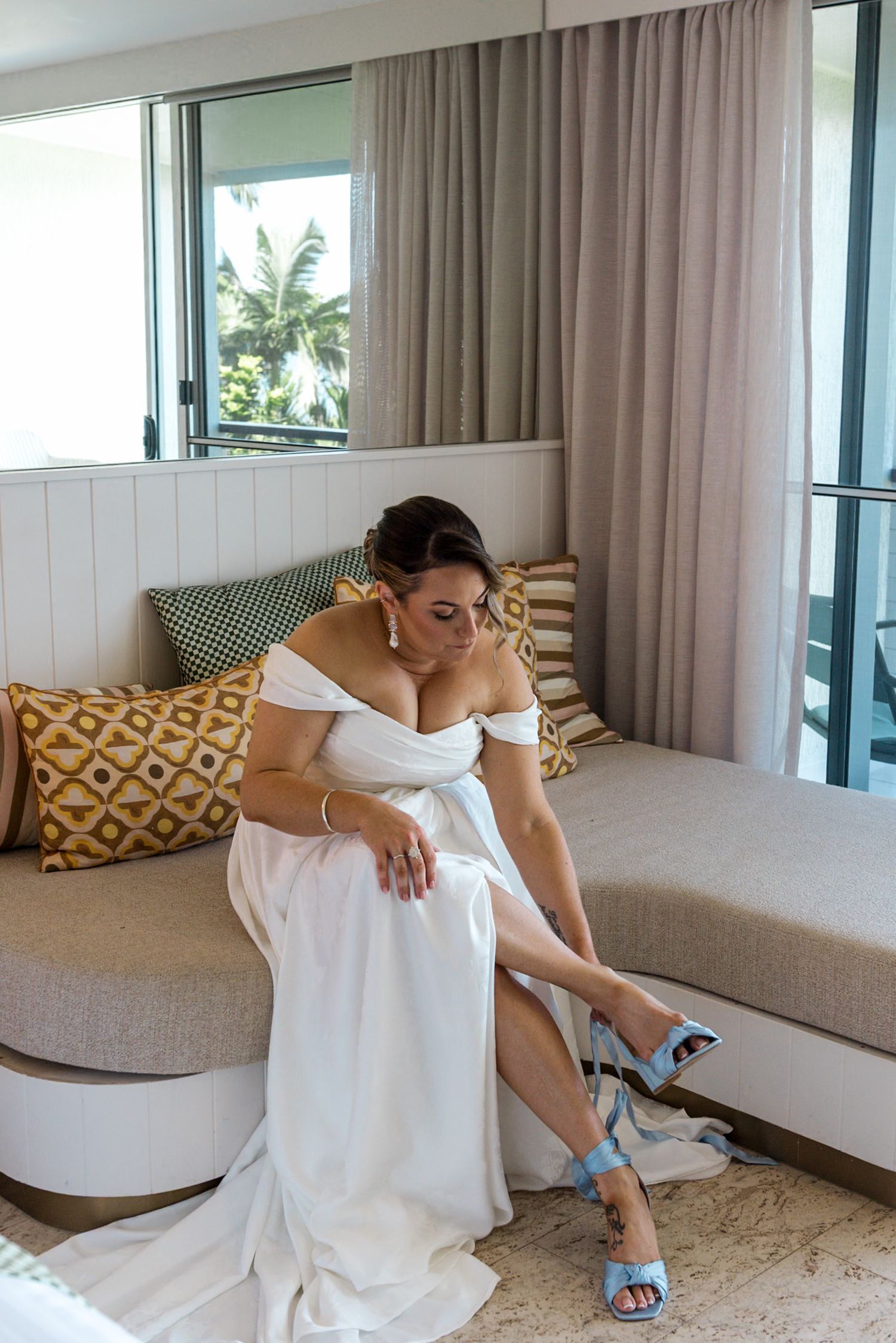 Bride in white off-shoulder gown fastening blue heeled sandals while seated on a cream sofa in a bright hotel room.