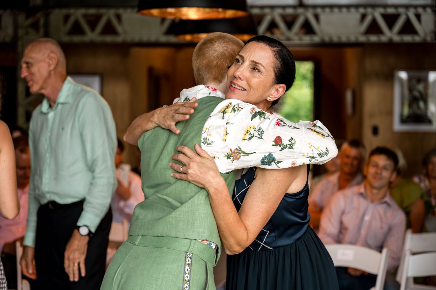 Two women share a warm hug inside a rustic wedding reception venue.