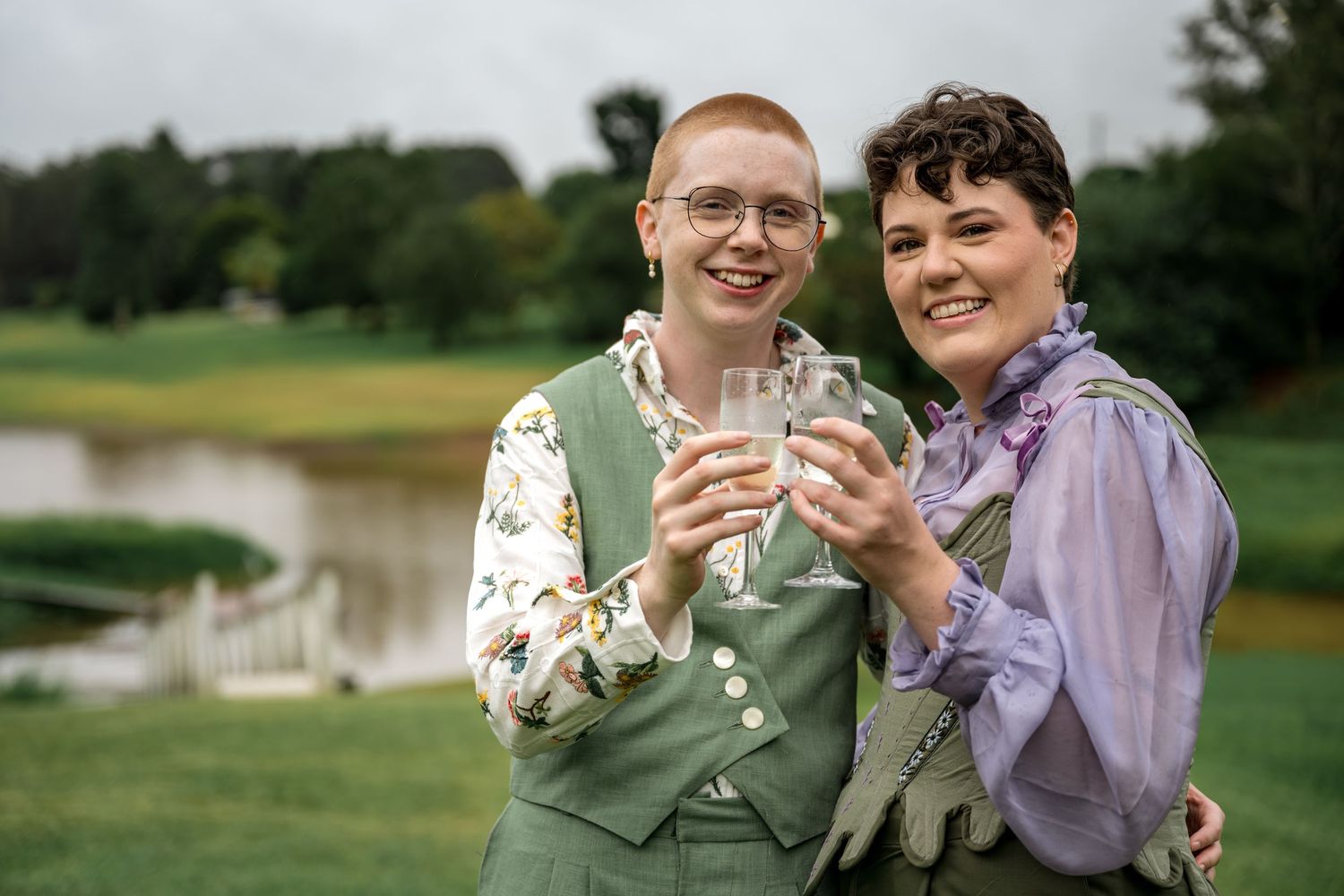 Two people in green outfits toasting with champagne glasses outdoors near a pond.