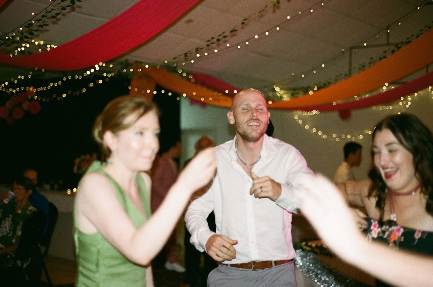 Man in white shirt dancing and smiling with two women at a festive wedding reception.