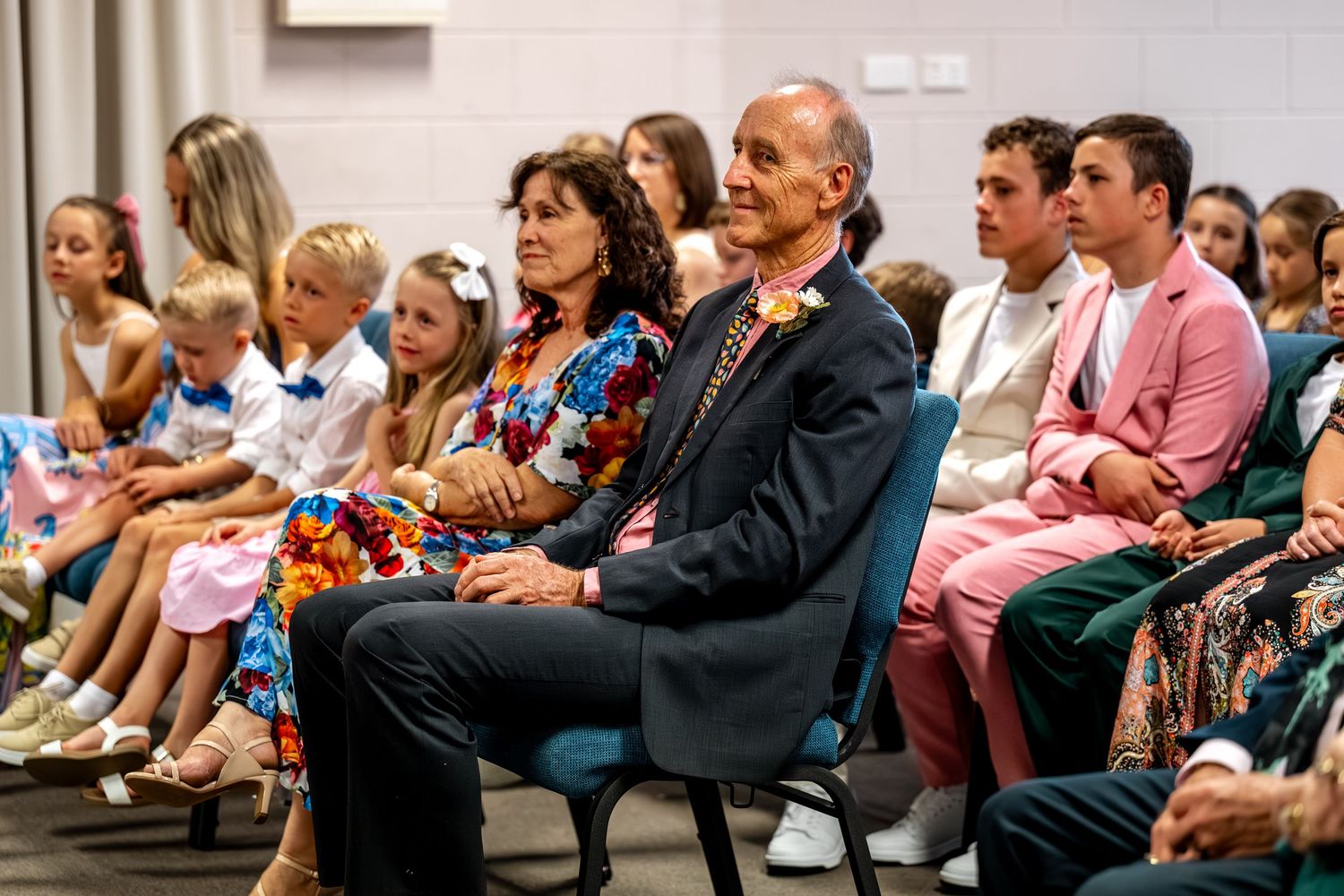 Wedding guests seated in rows, including a man in a navy suit with a floral boutonniere and woman in colorful dress.