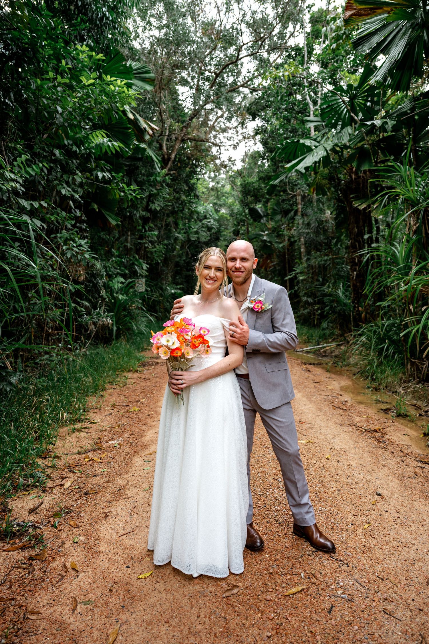 Wedding couple pose together on a tropical dirt road, groom embracing bride holding colorful orange bouquet.