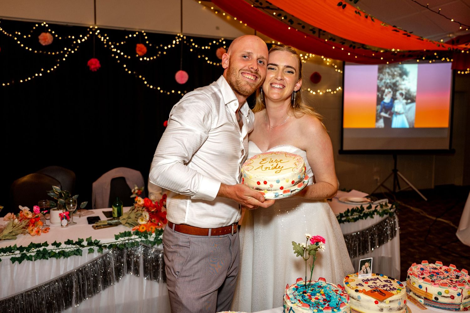 Happy bride and groom pose together holding a wedding cake at reception.