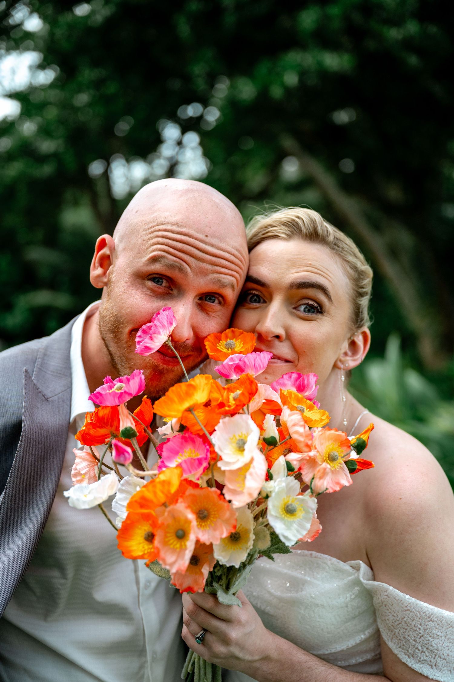 Playful bride and groom peeking over vibrant orange and pink poppy wedding bouquet with wide eyes and smiles.