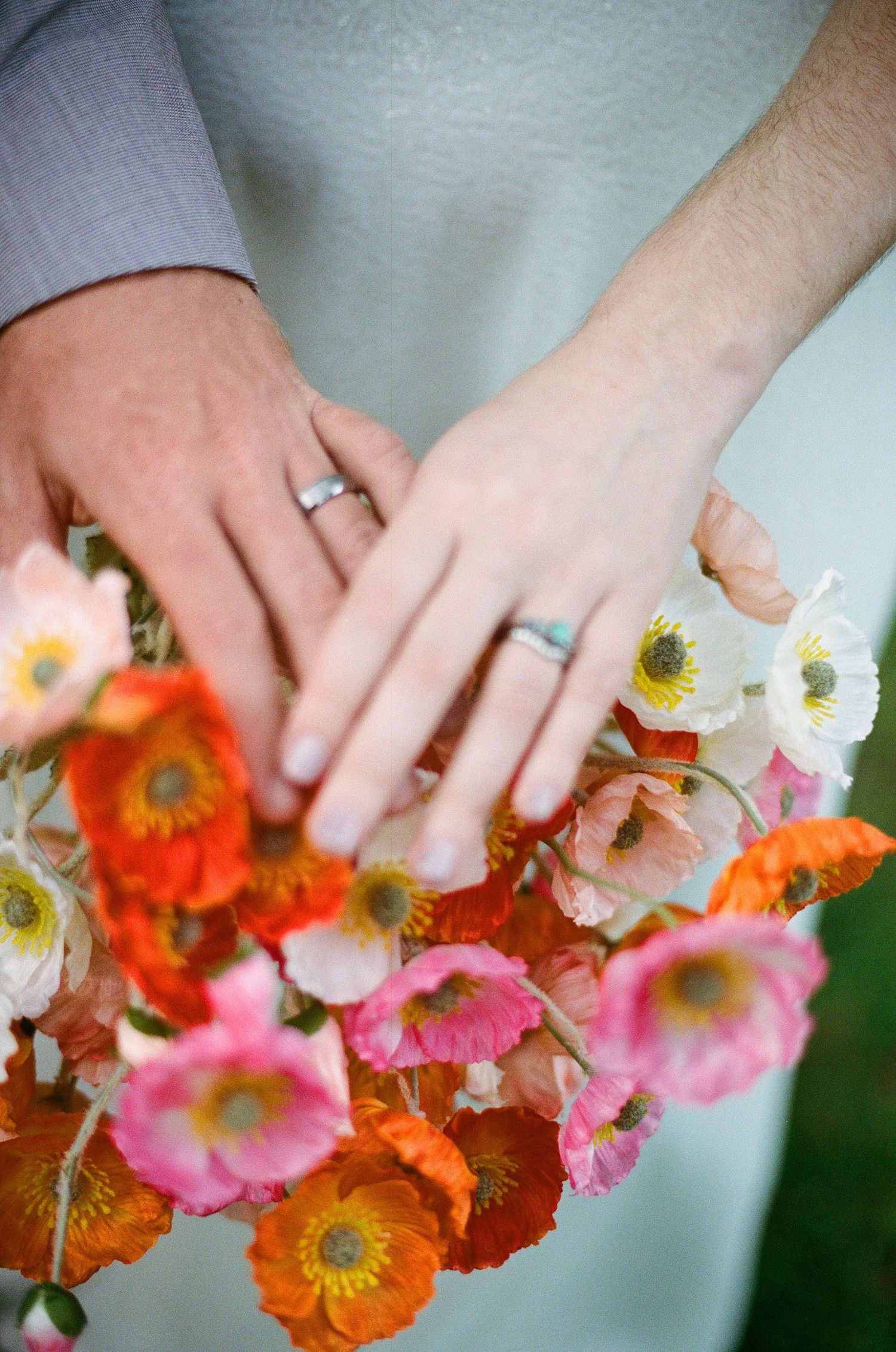 Newlyweds display wedding rings resting on a vibrant bouquet of colorful orange, pink, and white poppy flowers.