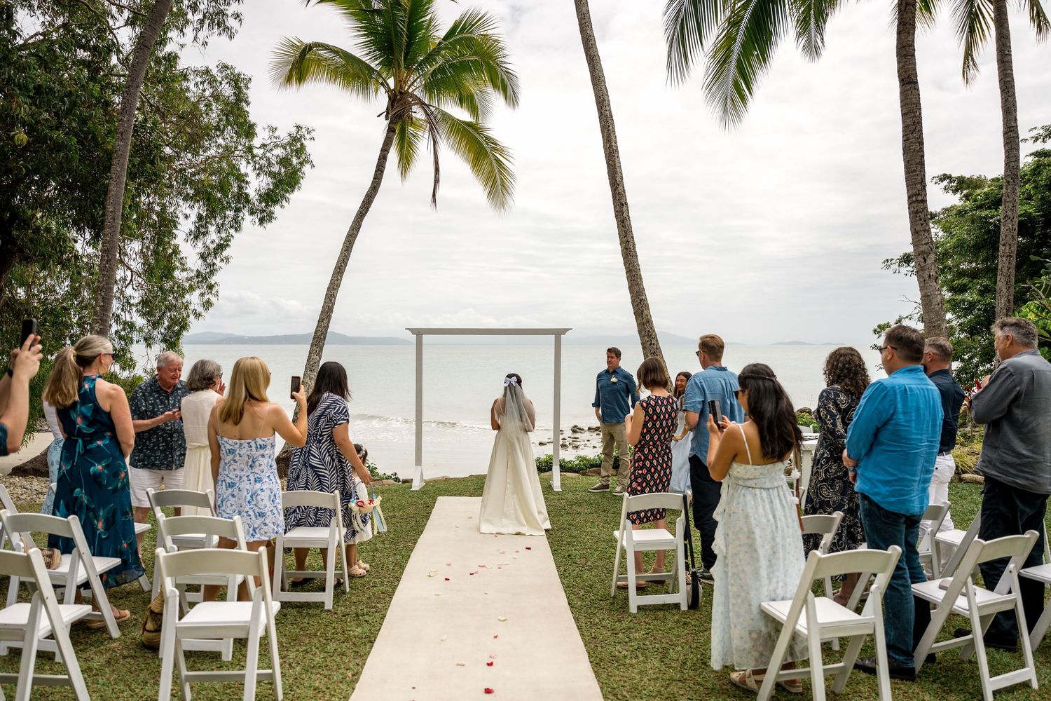 Outdoor beach wedding ceremony with white chairs and palm trees overlooking the ocean at Little Cove, Port Douglas.