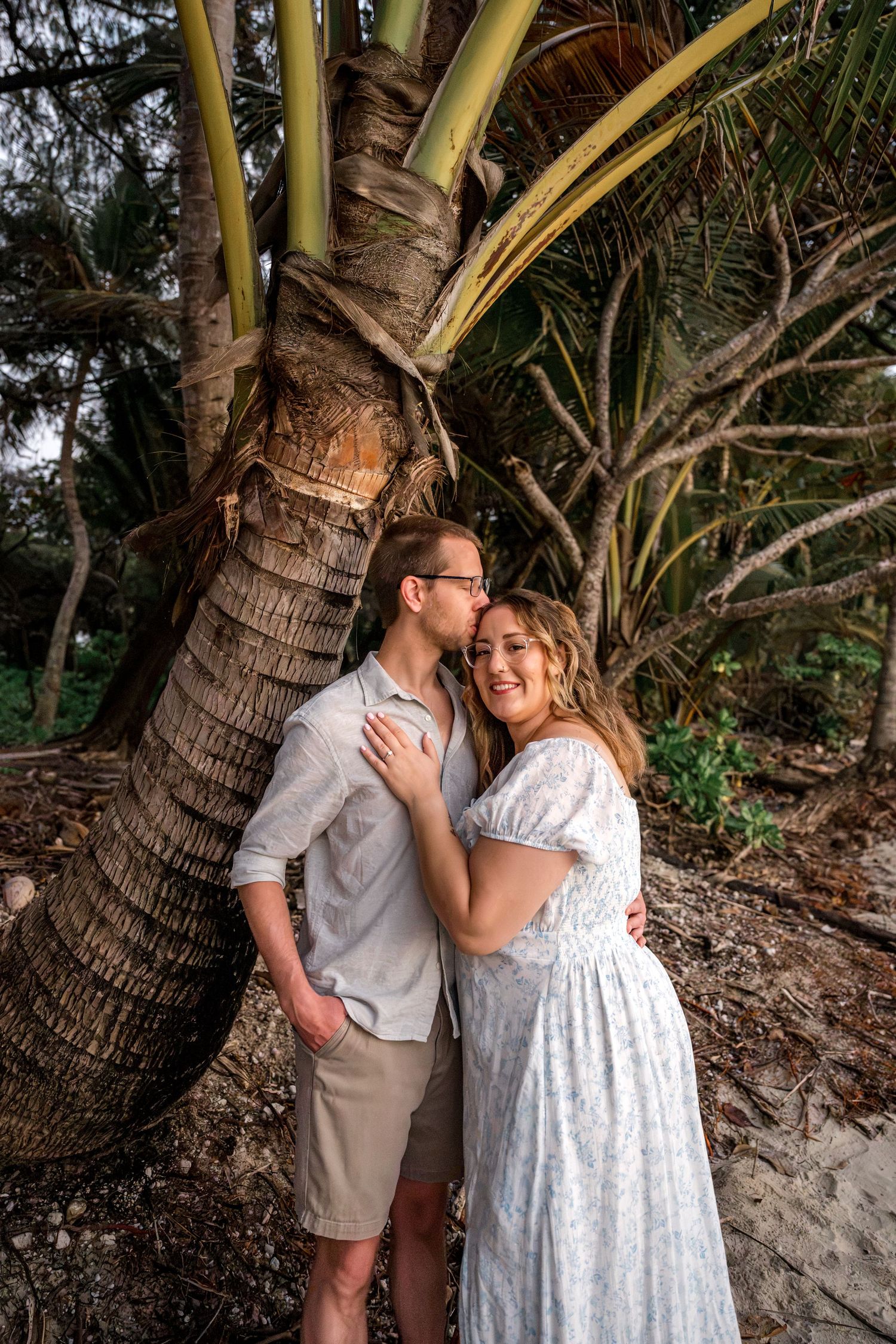 Man kissing woman on cheek as they embrace against a palm tree trunk on a tropical beach at Four Mile Beach, Port Douglas.