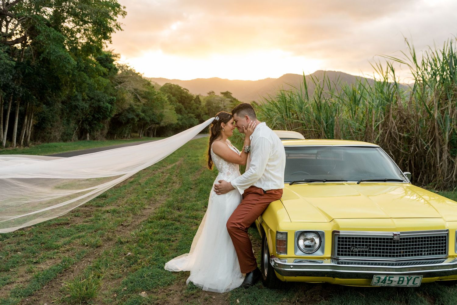 Bride and groom embrace beside a classic yellow car with veil blowing at sunset.
