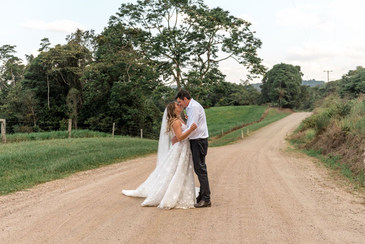 Bride and groom sharing a kiss on a rustic dirt road surrounded by lush tropical greenery.