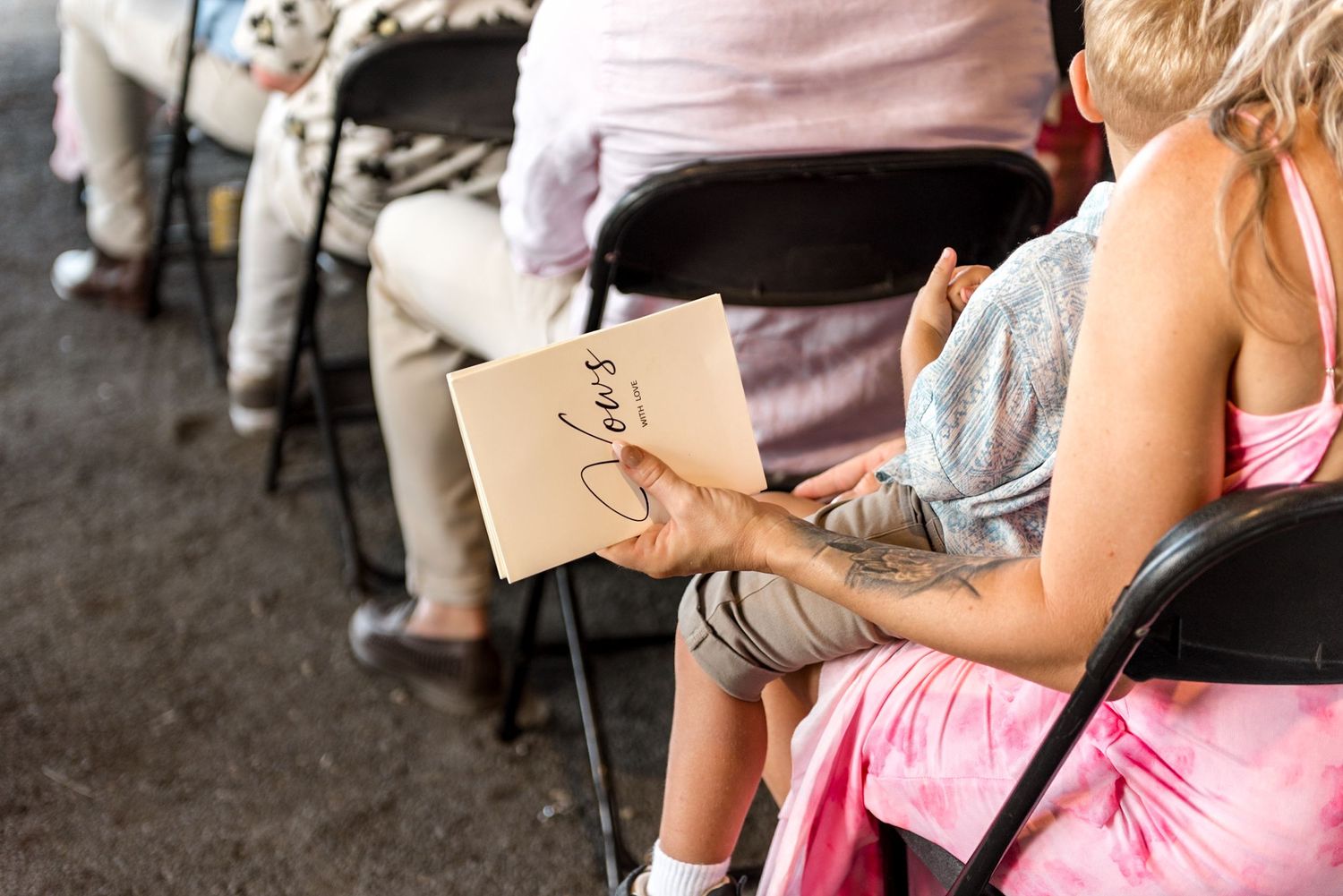 Wedding guest holds a vows booklet while seated with a child during an outdoor barn wedding ceremony.