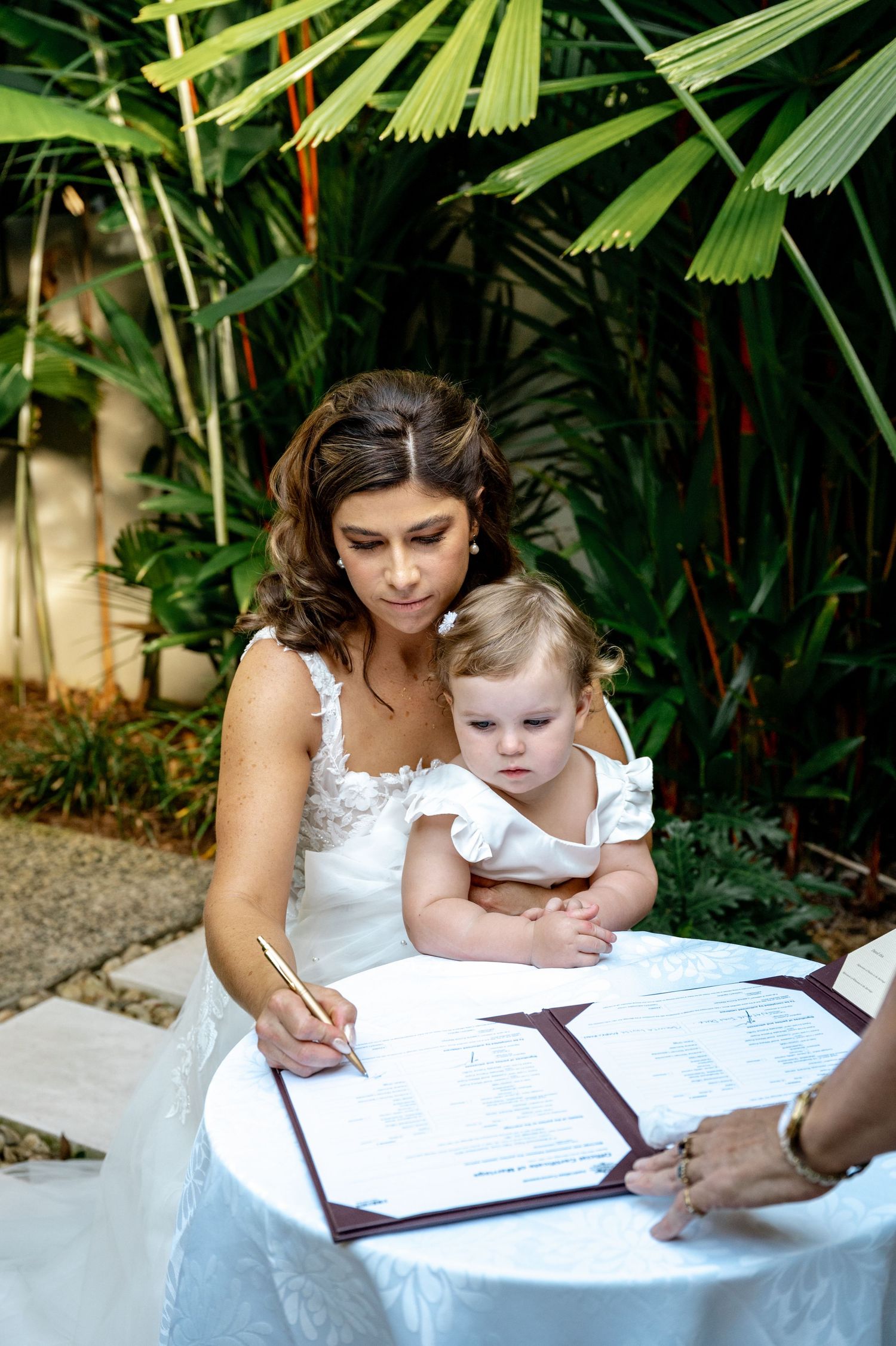 Bride in white lace gown signing marriage certificate while holding baby at tropical outdoor ceremony.