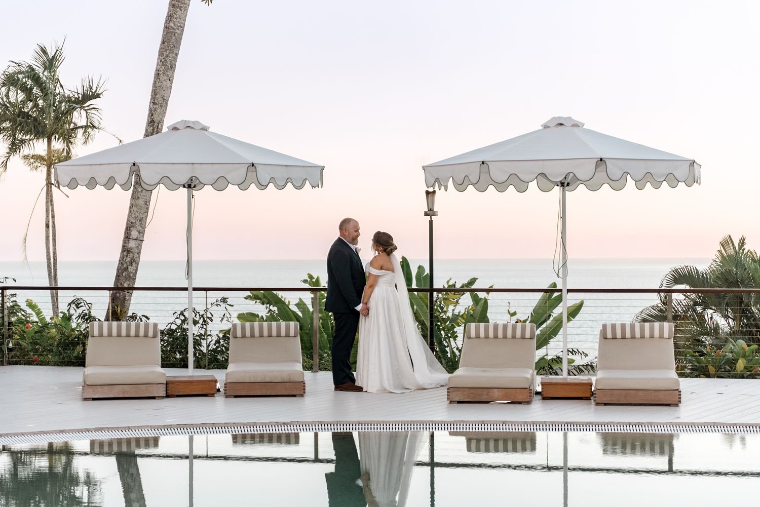Bride and groom stand together by a luxury pool with white umbrellas and stunning ocean sunset views.