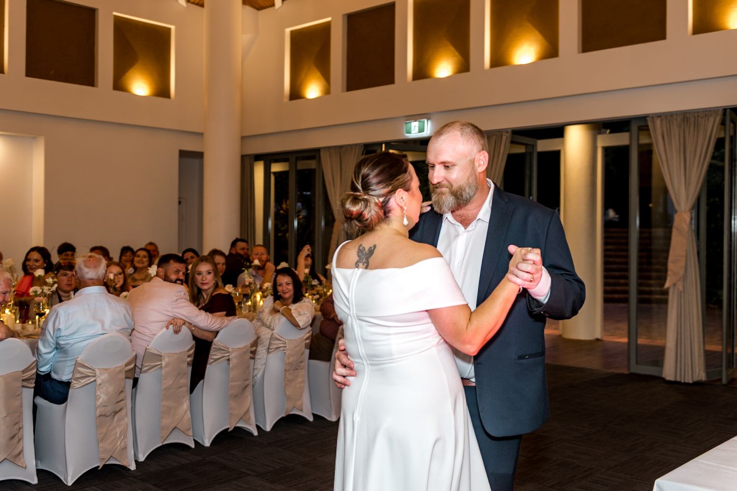 Bride in white off-shoulder gown and groom in navy suit share intimate first dance at wedding.