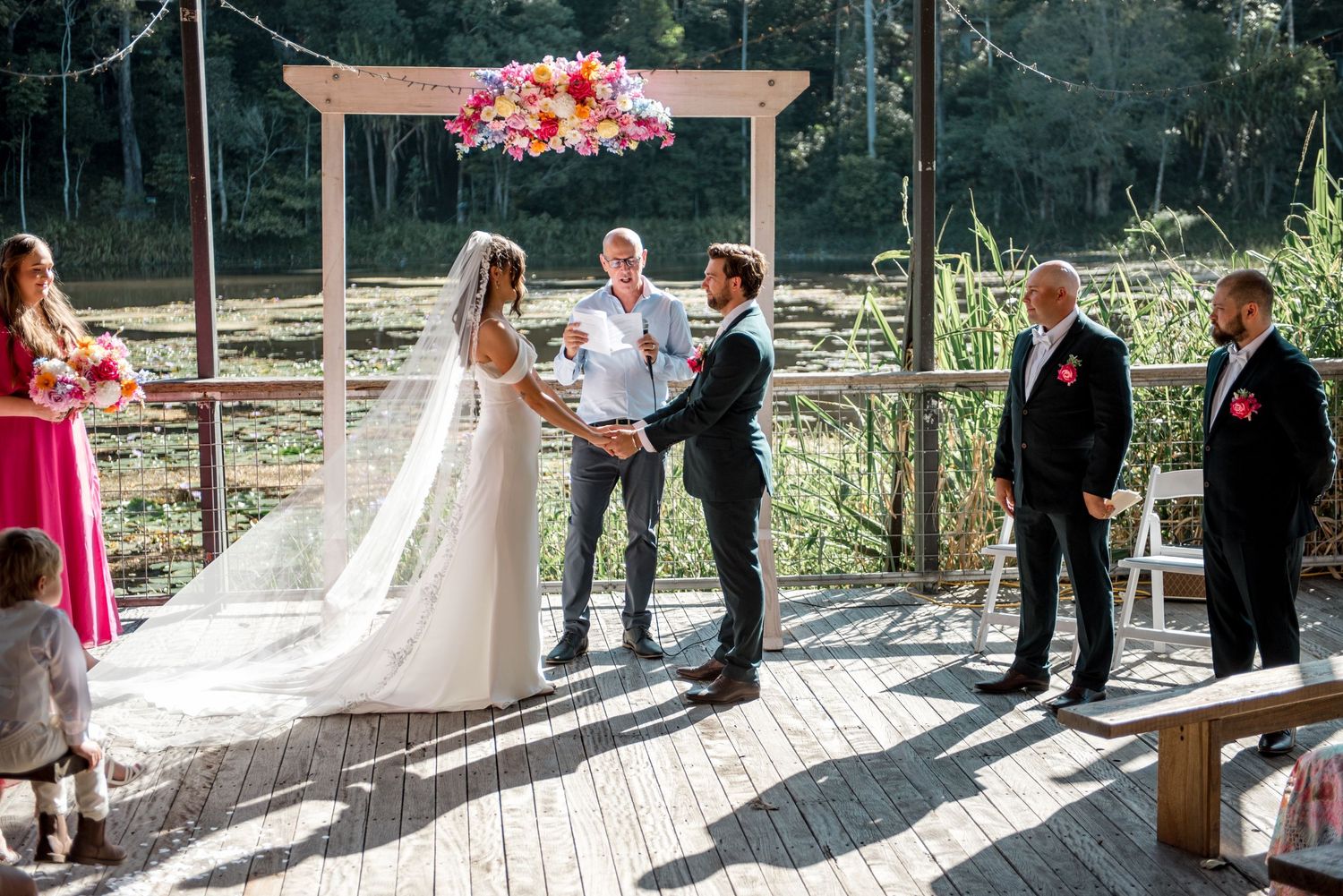 Bride and groom exchange vows at outdoor waterfront ceremony under floral arch with wedding party.