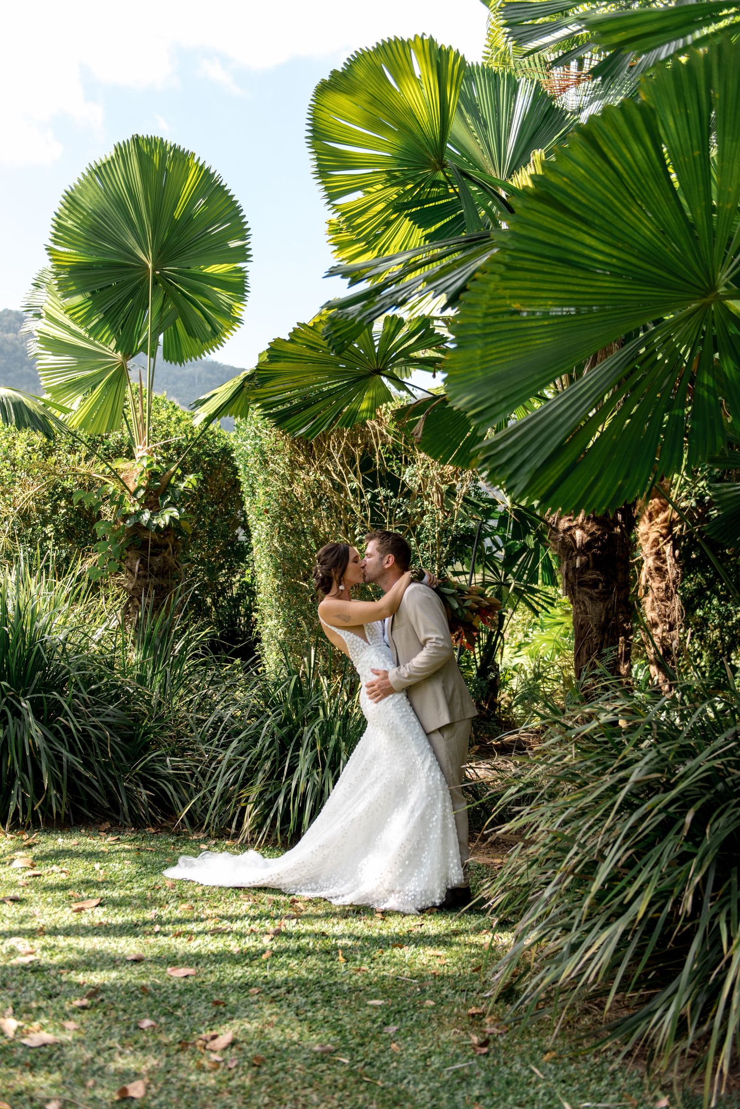 Bride and groom kissing under lush tropical fan palm trees in a garden wedding setting.