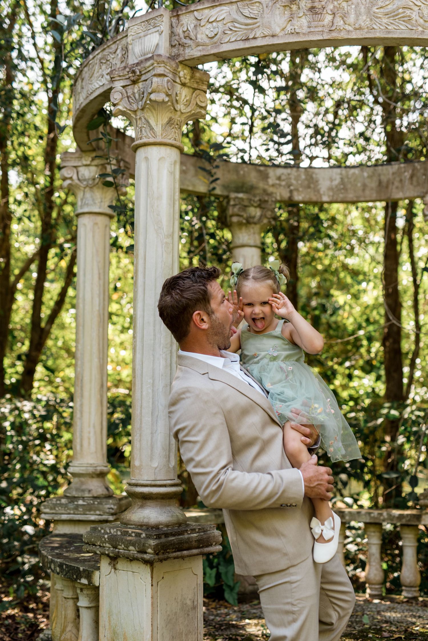Groom in tan suit laughing with young girl in green dress beside ornate stone gazebo in garden.