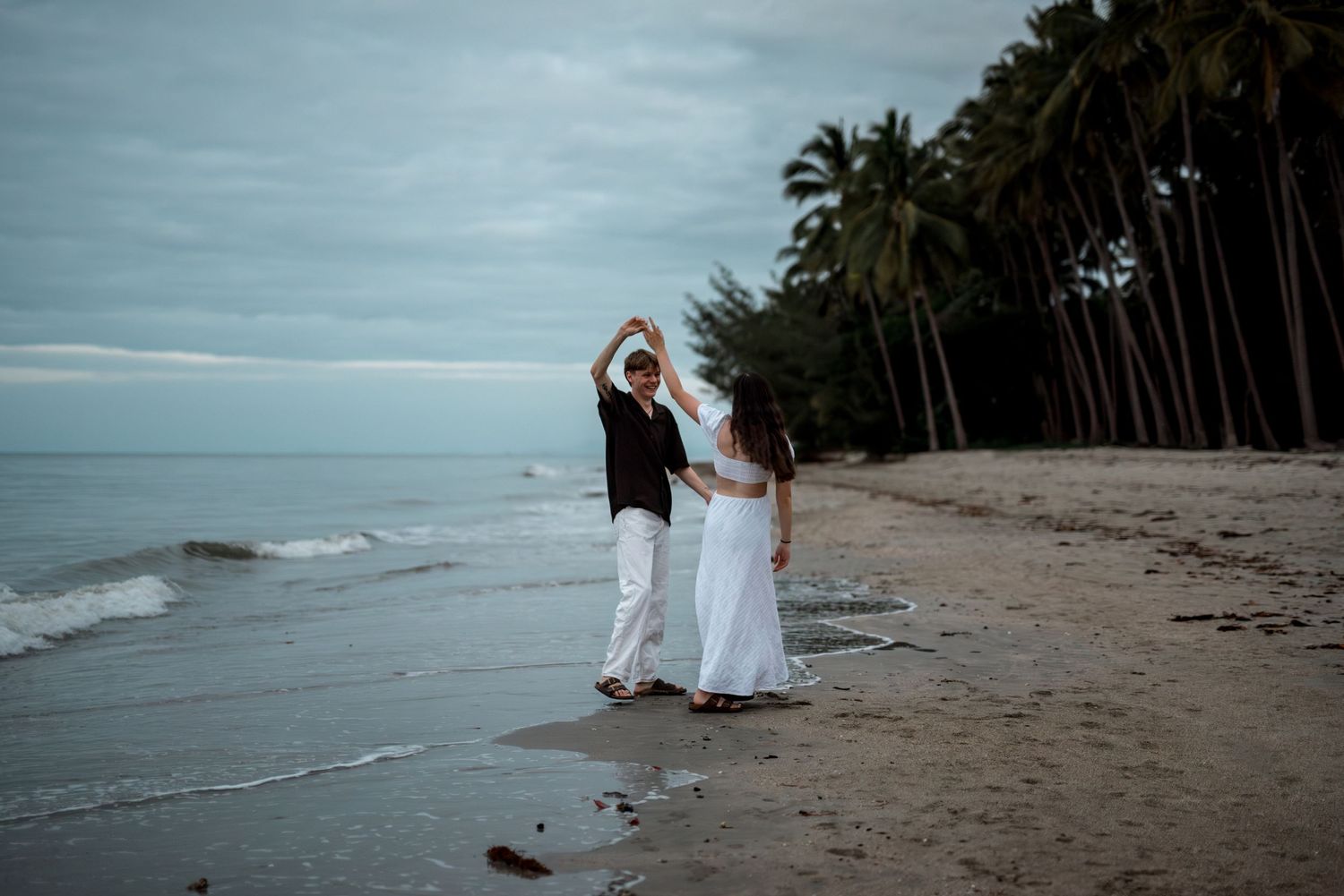 Couple dancing on a tropical beach at dusk with palm trees and calm ocean waves nearby.