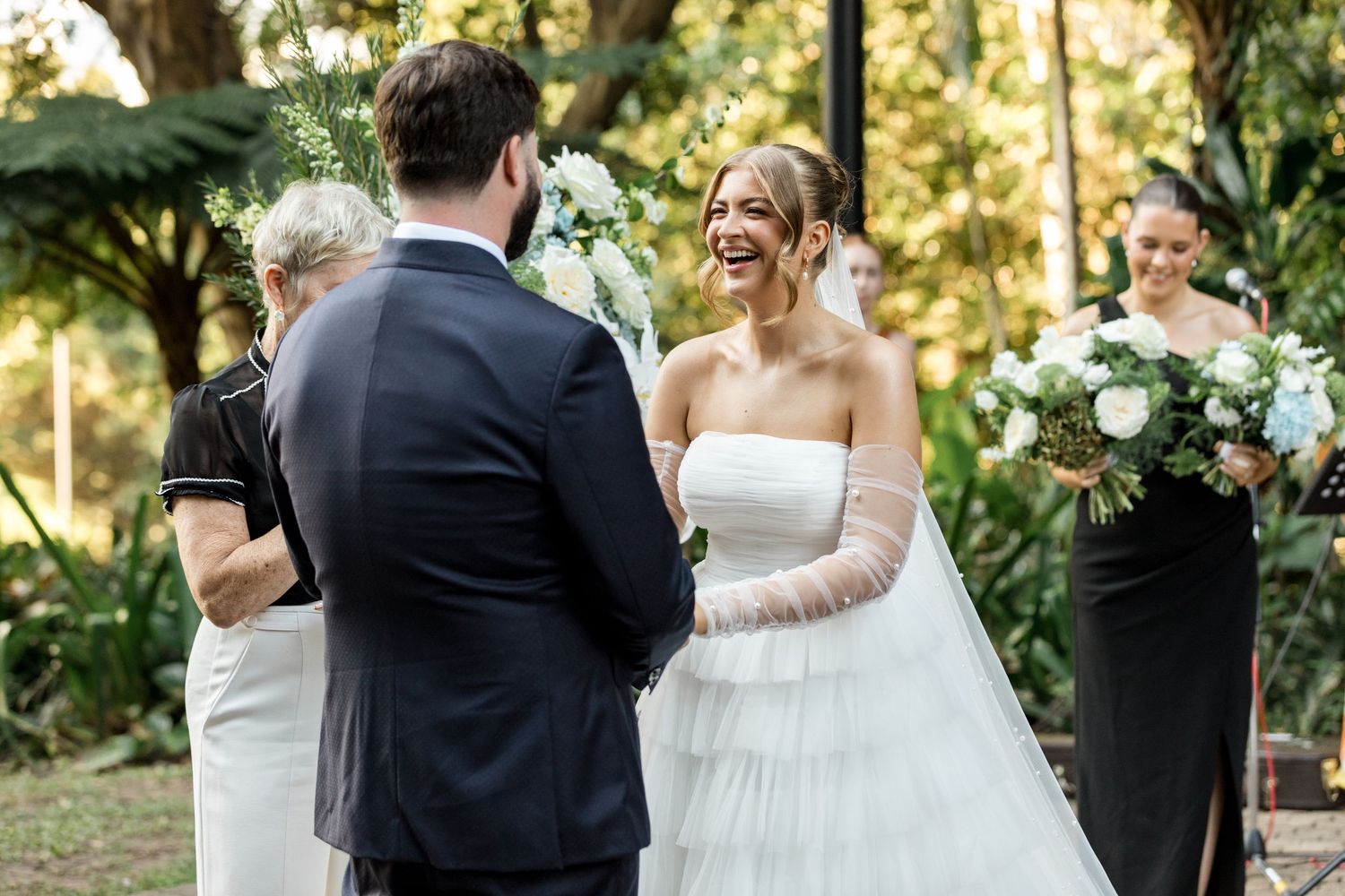 Bride in white strapless gown laughs with groom as bridesmaid in black dress stands nearby.