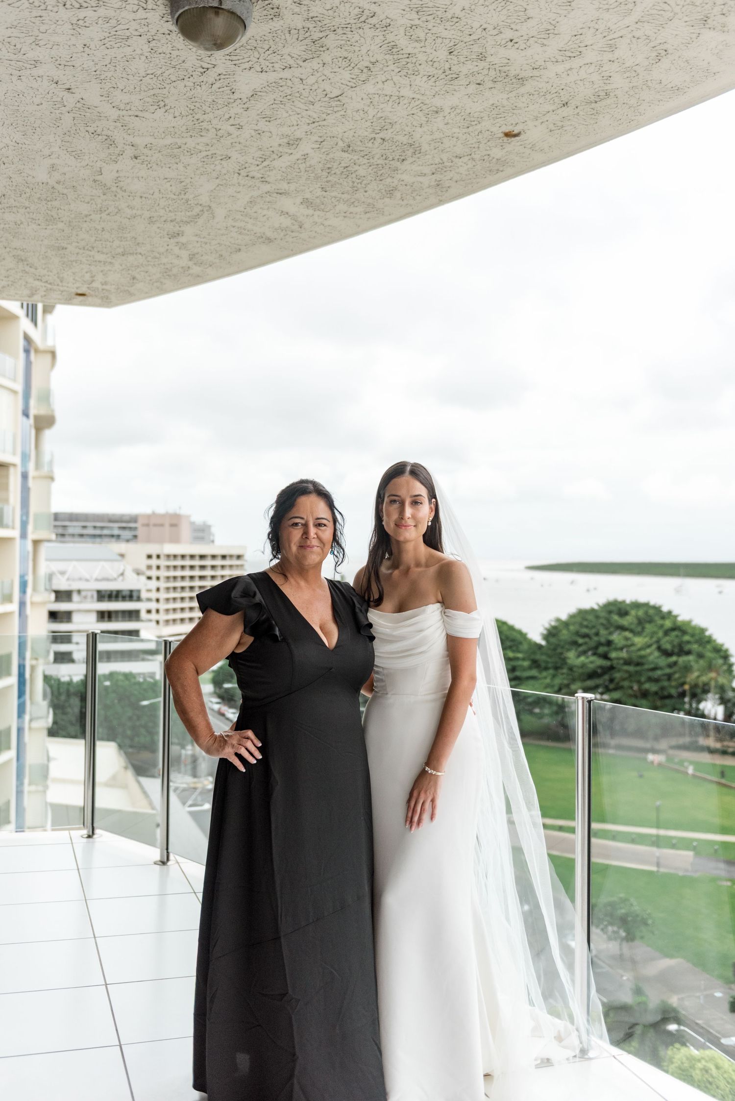 Bride in white off-shoulder gown and veil poses with mother in black dress on high-rise balcony.
