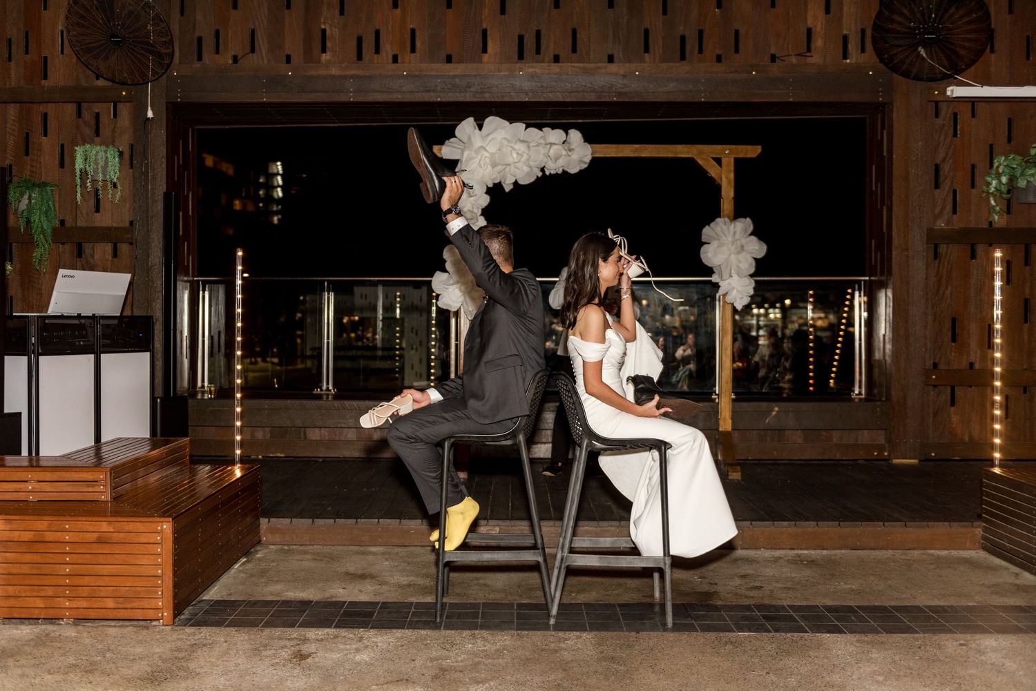 Bride and groom sit back-to-back on chairs at night with balloon arch decor behind them.
