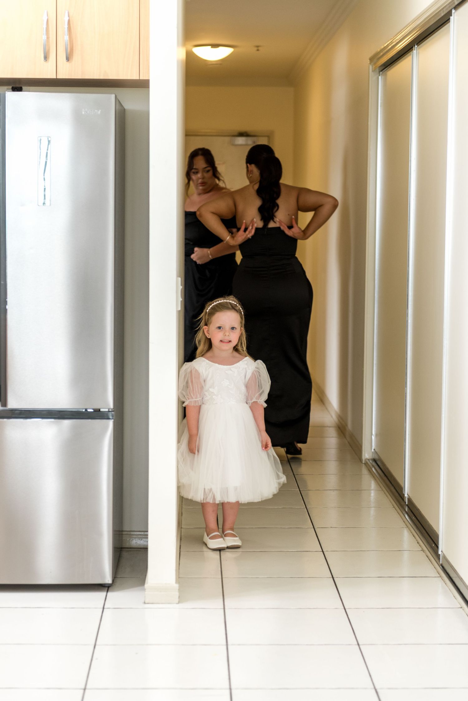 A smiling flower girl in a white dress peeks around a corner while bridesmaids in black dresses prepare in the background.