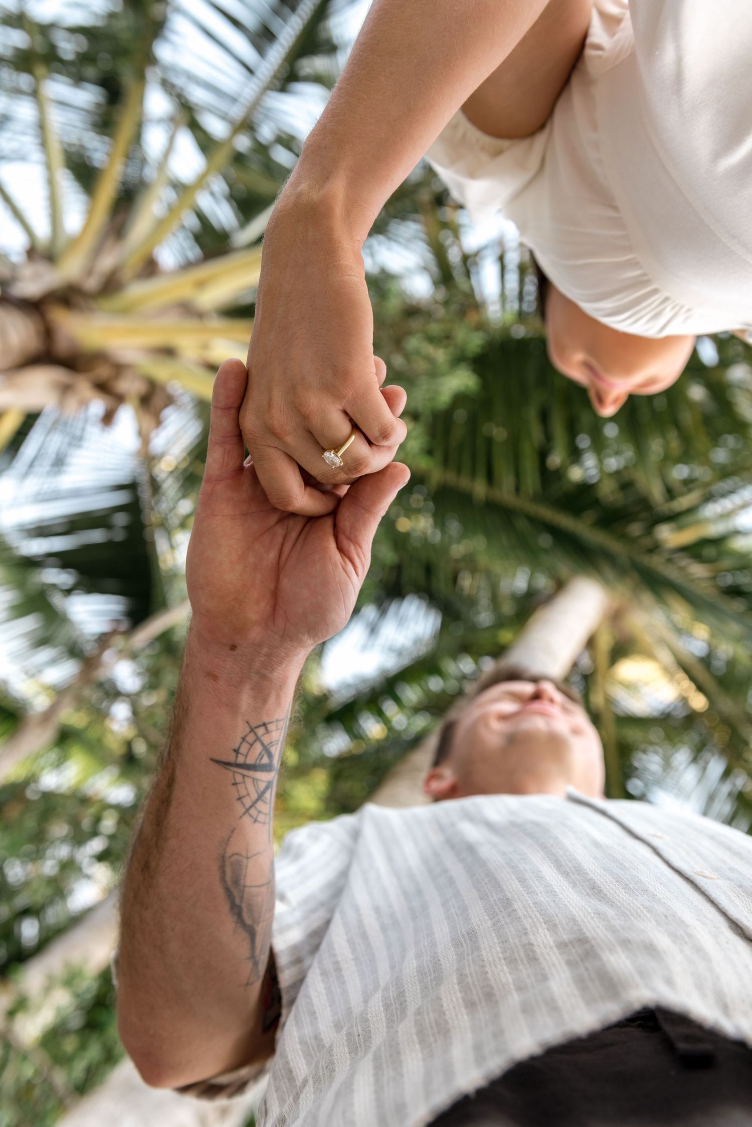 Low-angle view of a couple holding hands beneath a tropical palm tree, showcasing a ring and tattoo.