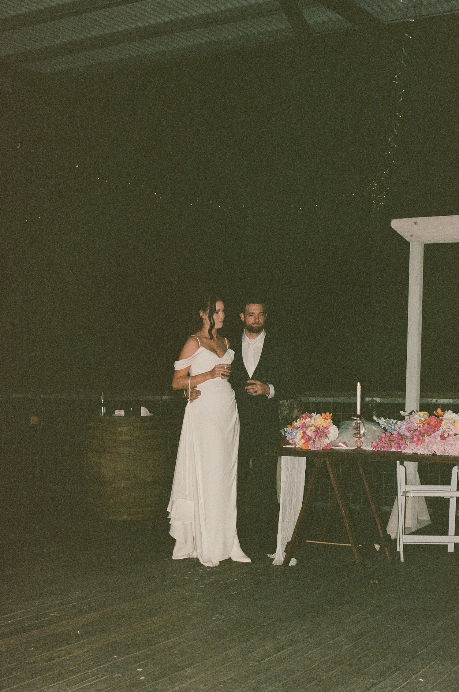 Bride and groom standing together at night near floral wedding table at barn venue.
