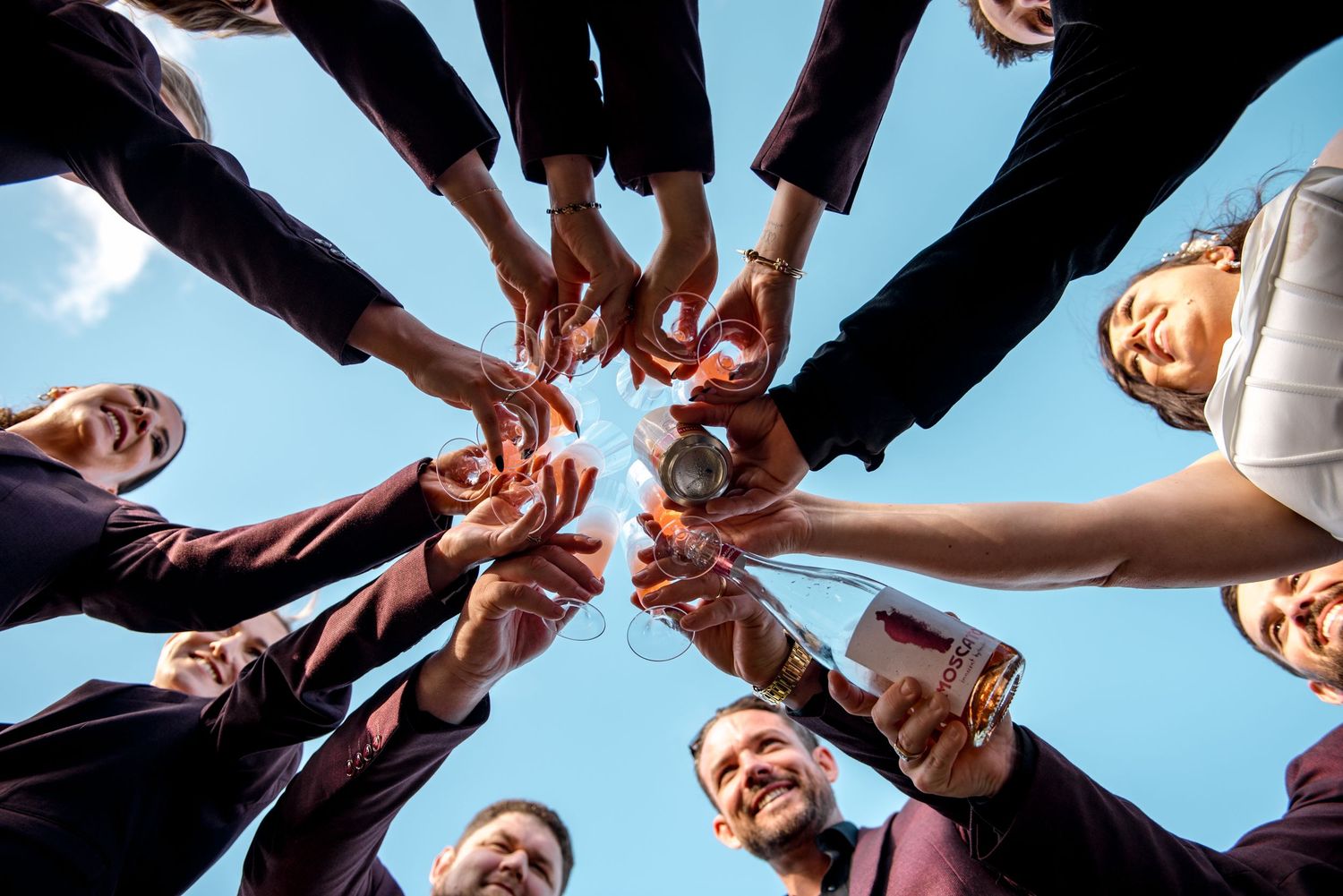 Wedding party clinking glasses in a circle viewed from below against blue sky.