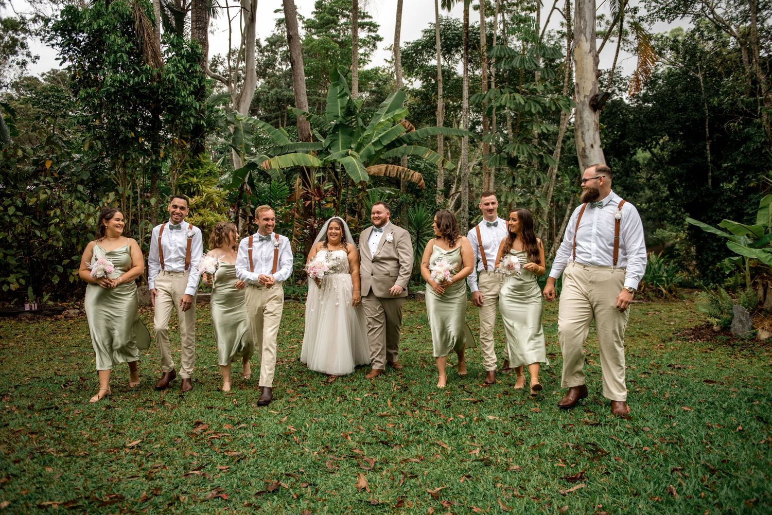 Full wedding party walks together outdoors in sage green and tan attire with tropical greenery backdrop.