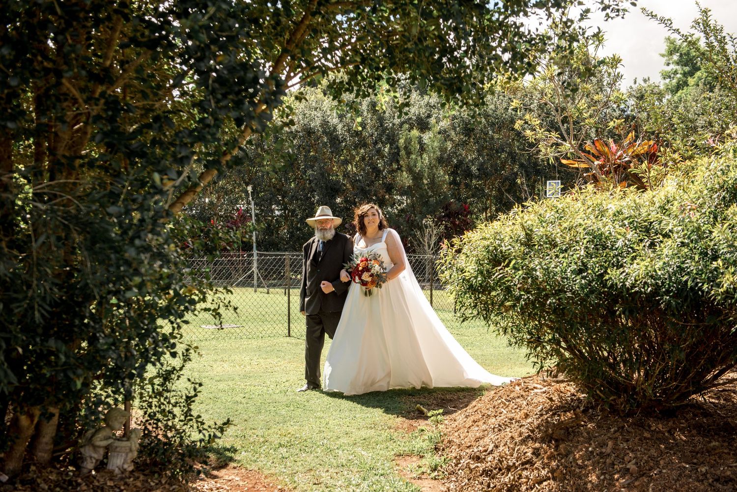 Bride in white gown holding autumn bouquet walks with older man in hat through garden path.