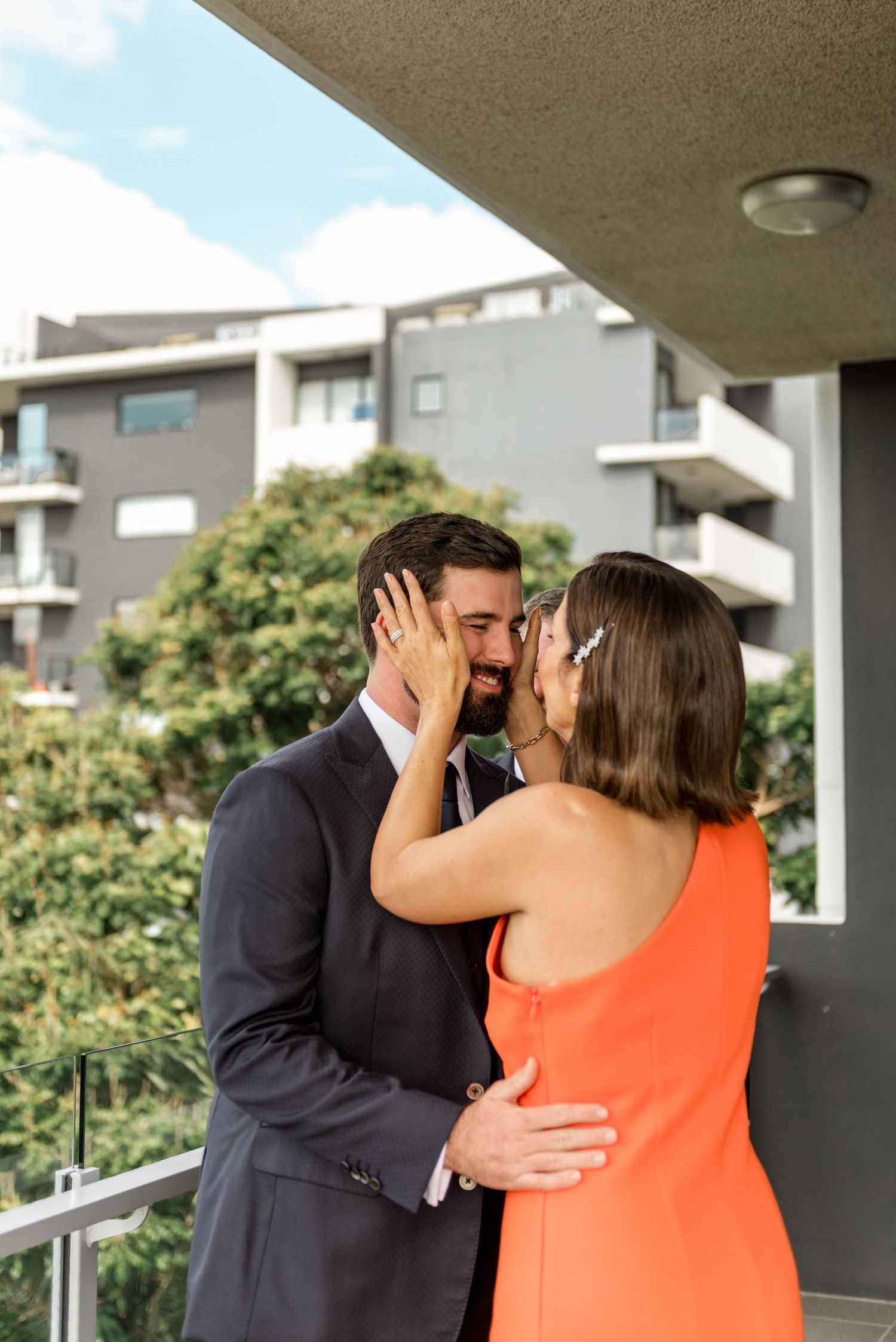 Groom in dark suit shares a tender kiss with woman in orange dress on a balcony with apartments in background.