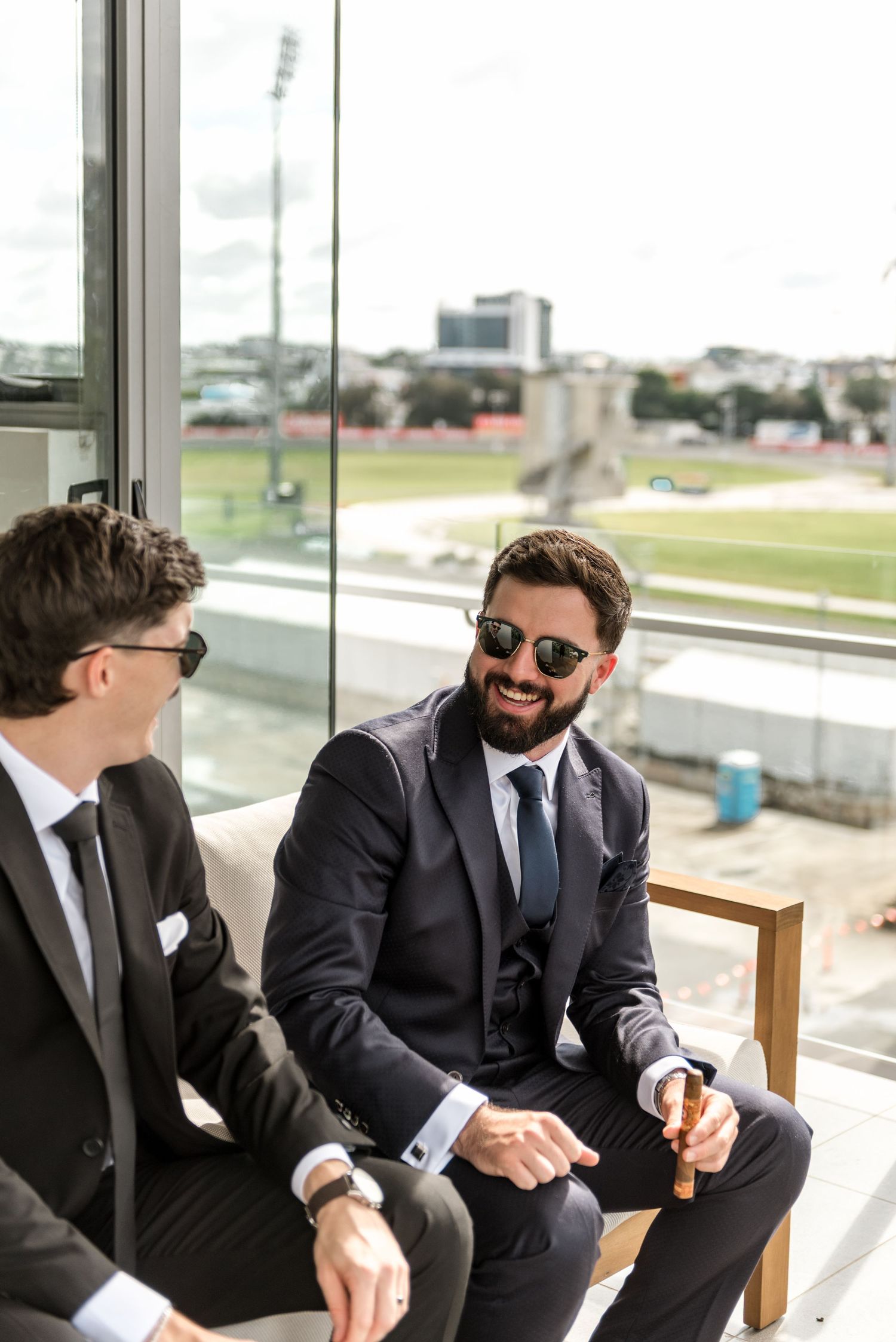 Two groomsmen in black suits and sunglasses share a laugh while relaxing near a window overlooking a sports stadium.