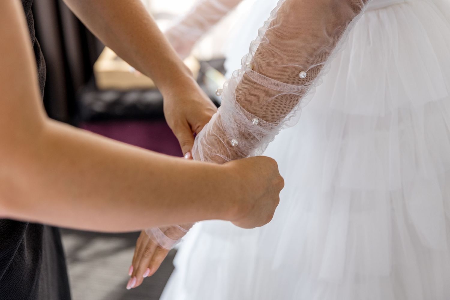 Bridesmaid fastening sheer pearl-adorned tulle sleeve of brides layered white wedding dress.