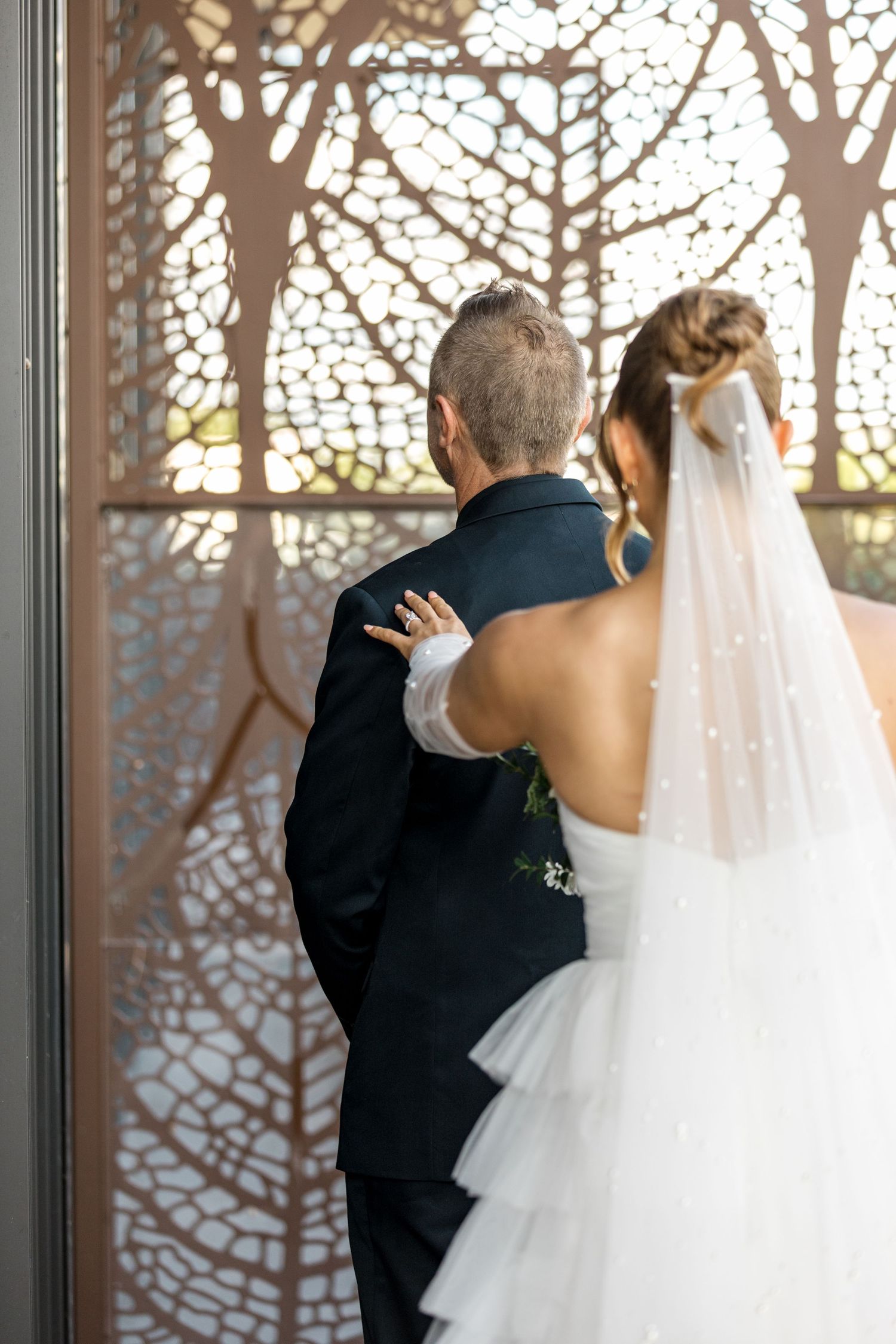 Bride in pearl veil places hand on groom's back as they face ornate laser-cut screen.