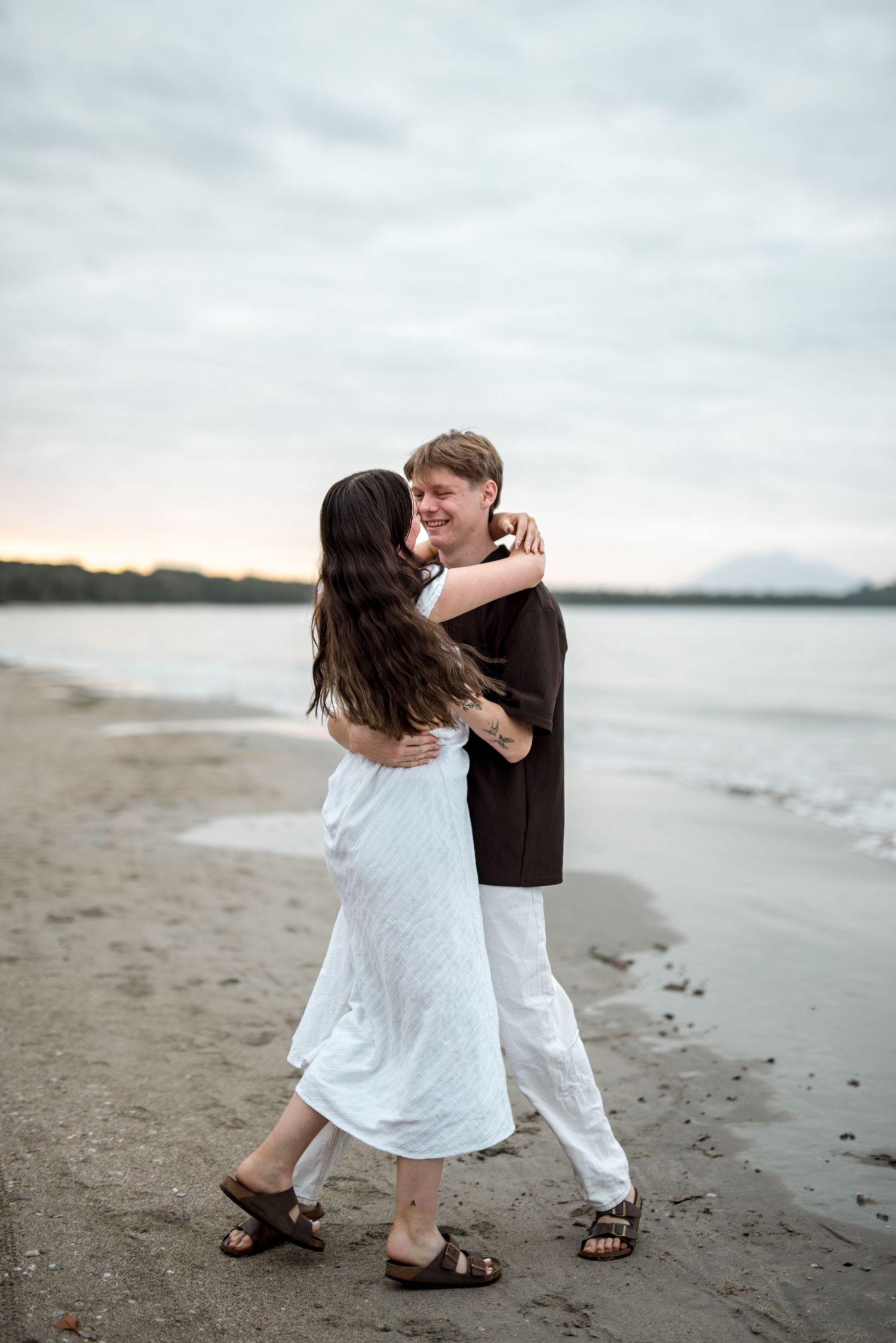 Laughing couple dances and embraces on sandy beach at overcast sunset.