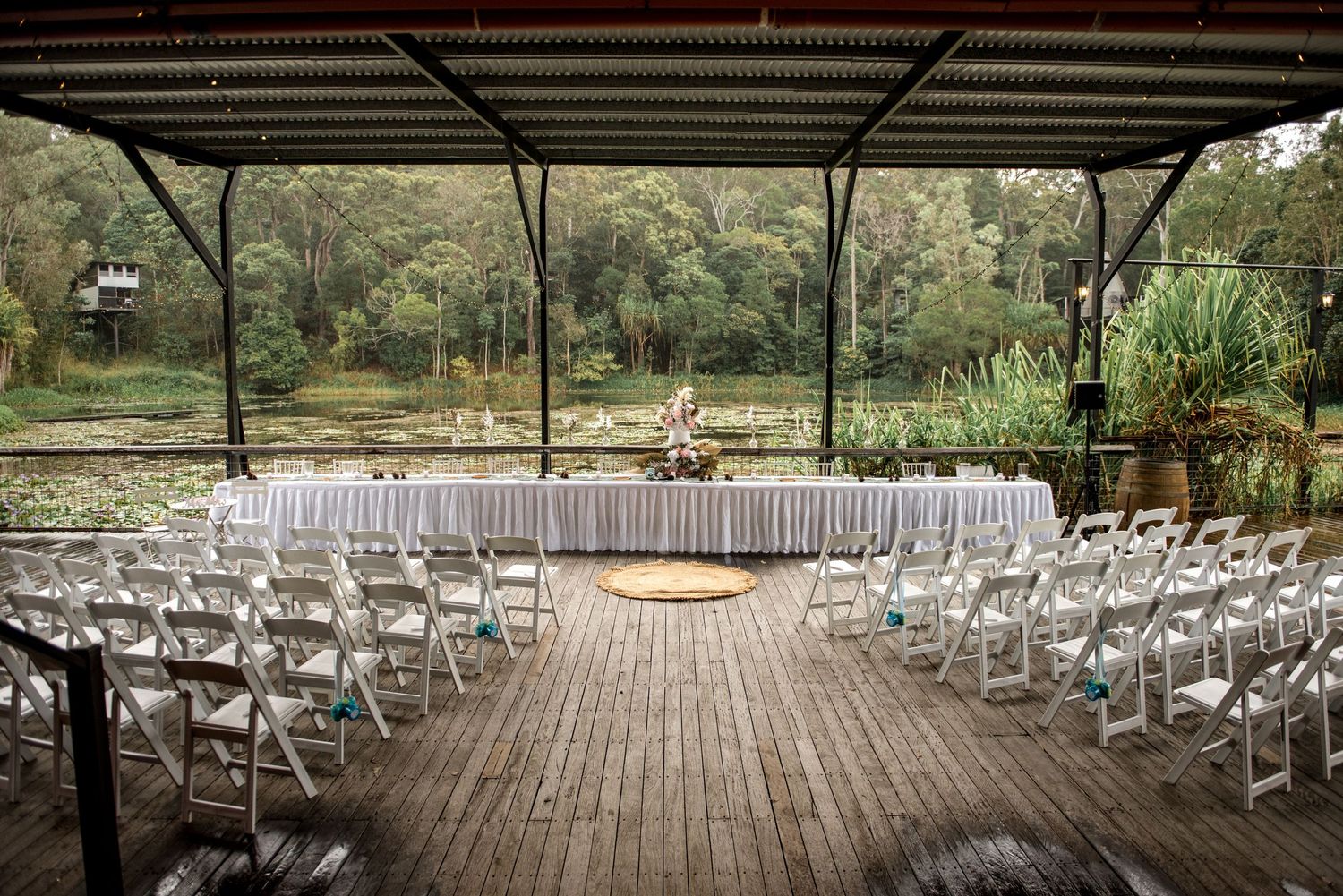 Outdoor wedding ceremony setup with white chairs and head table under a rustic pavilion by a pond.