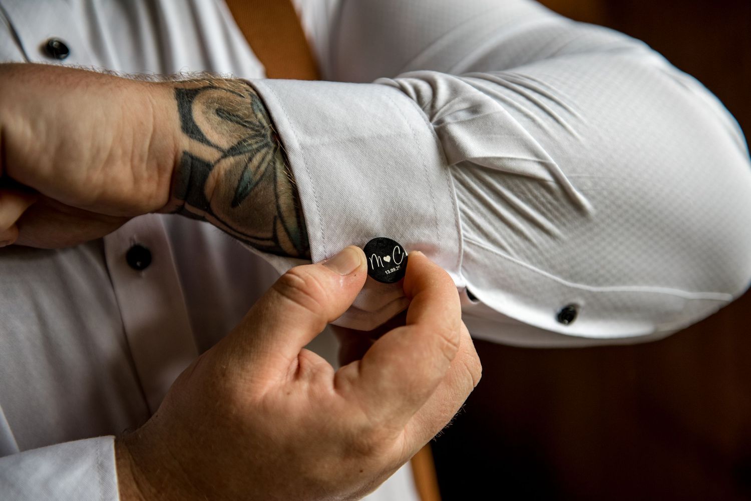Close-up of tattooed hands fastening a dark cufflink on a white dress shirt sleeve.