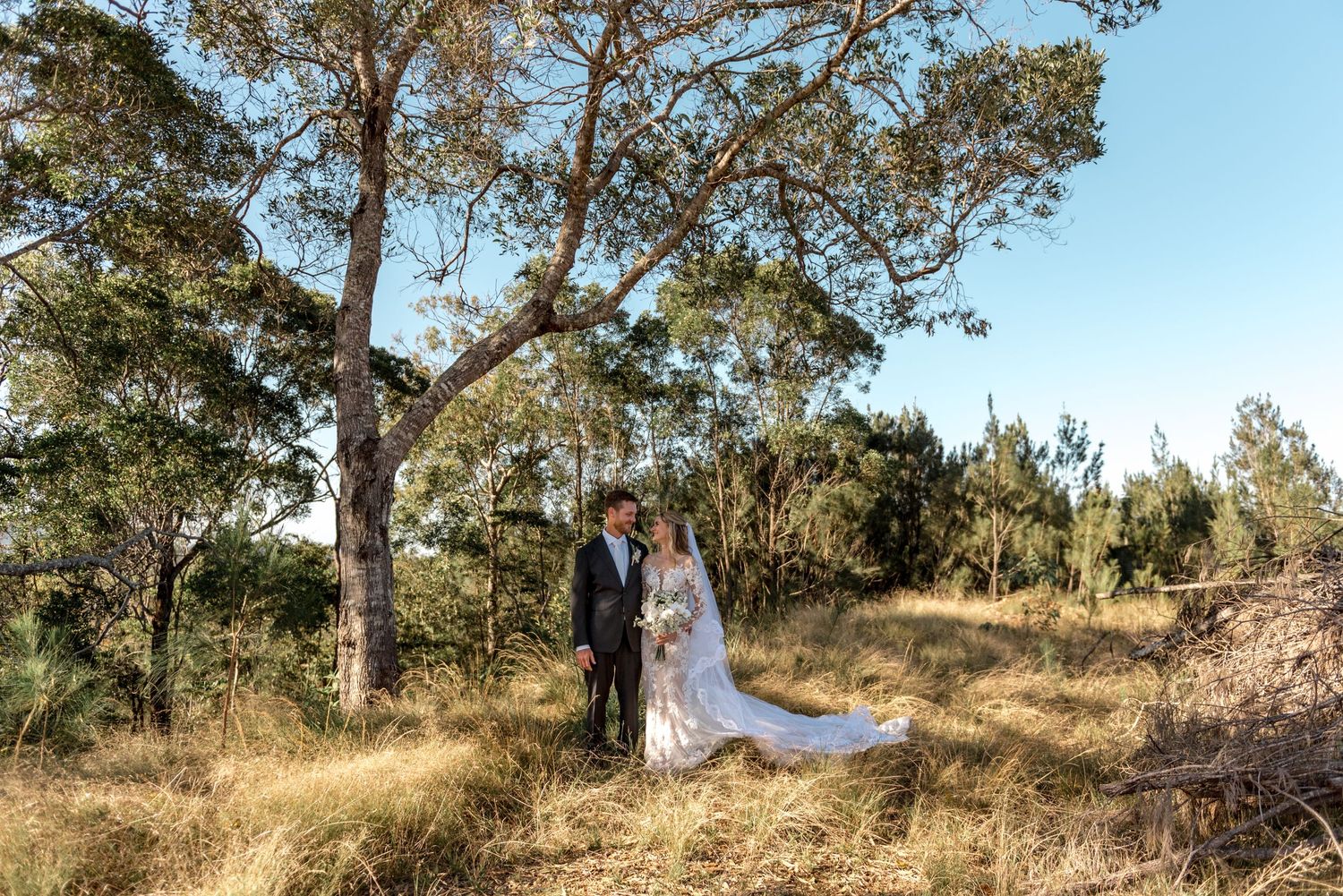 Bride and groom standing together under large tree in golden grass with blue sky behind.