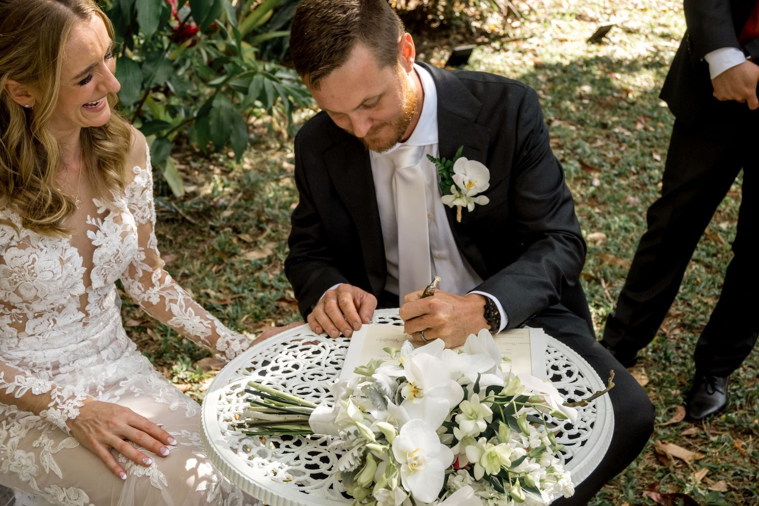 Groom signs wedding certificate as smiling bride watches at outdoor garden ceremony.
