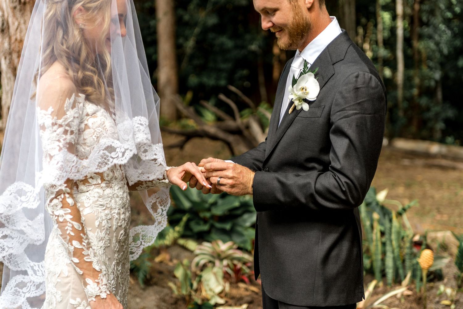 Groom placing wedding ring on bride's finger during outdoor forest ceremony.