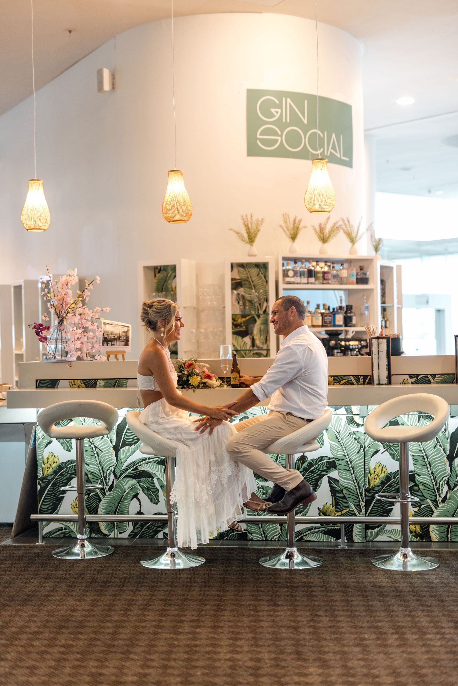Newlyweds hold hands and share an intimate moment seated at a tropical Gin Social bar.