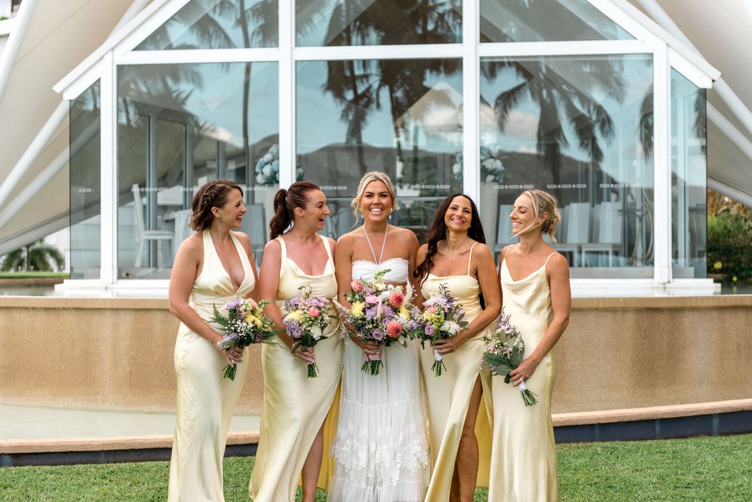 Bride poses with four bridesmaids in yellow satin dresses holding colorful bouquets at a chapel.