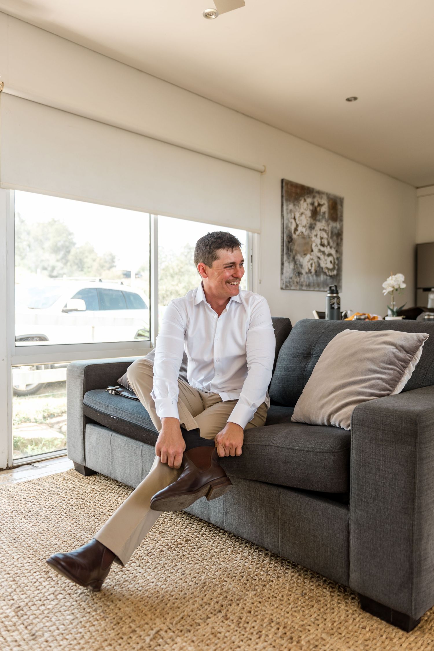 Smiling groom in white shirt and brown Chelsea boots sitting on grey couch during wedding preparations.
