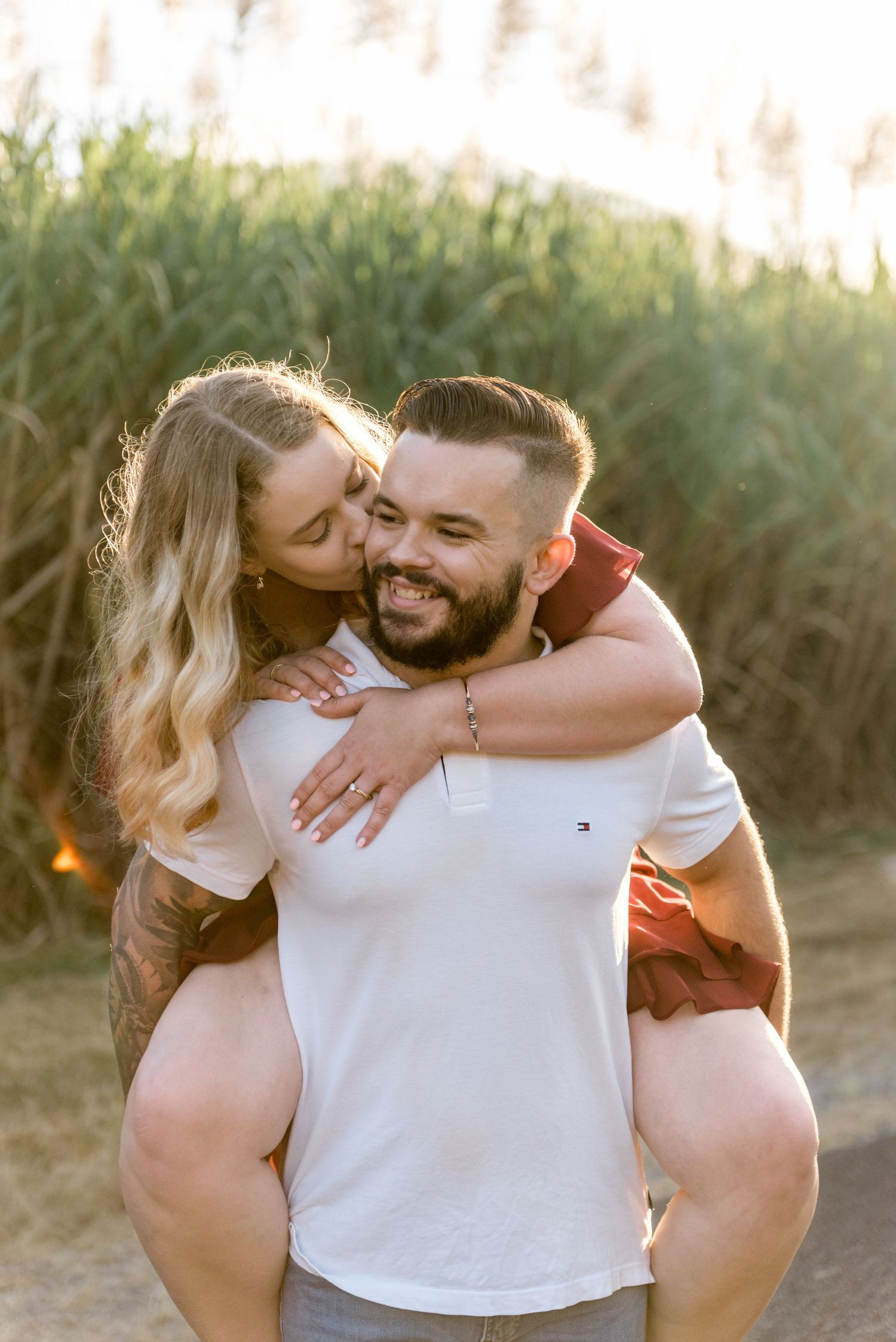 A woman kisses a smiling bearded man on the cheek during a sunny piggyback ride outdoors.
