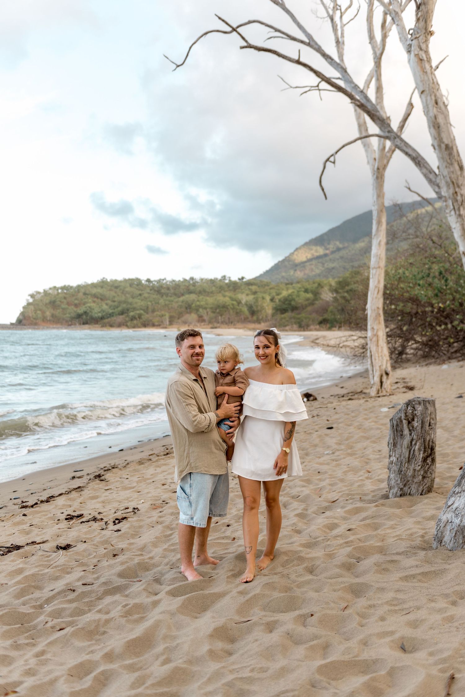 Happy family with toddler posing on tropical beach with ocean and green hills behind.