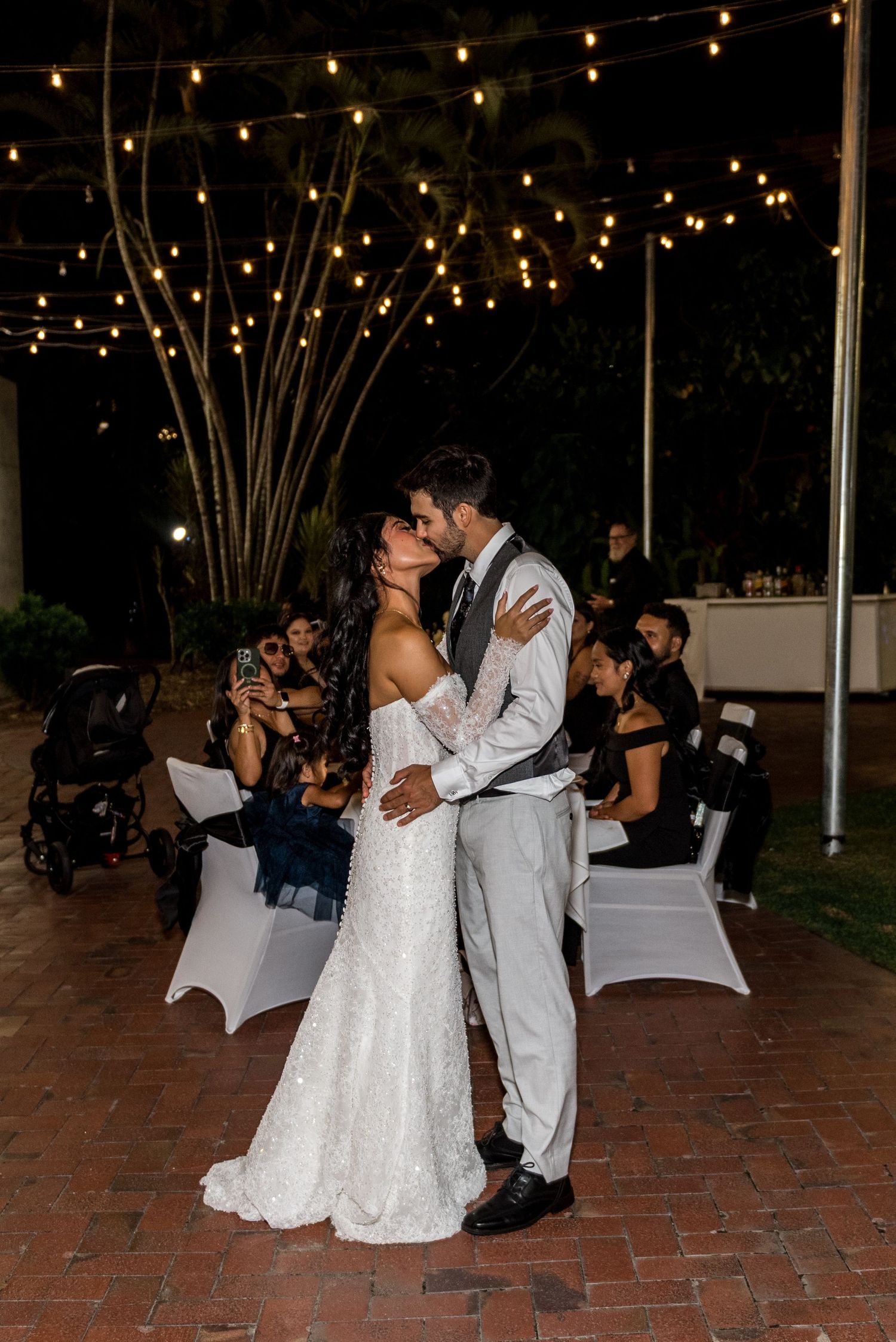 Bride and groom share a romantic kiss under warm string lights during their first dance at a night wedding reception.