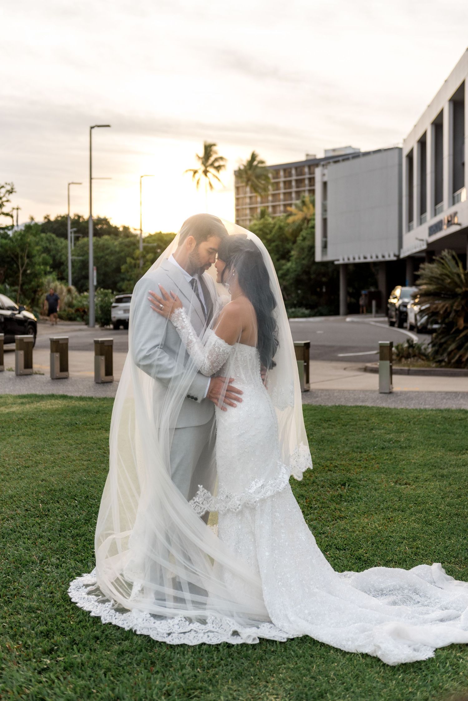 Newlyweds embrace under a flowing lace veil at golden sunset with soft warm backlight behind them.